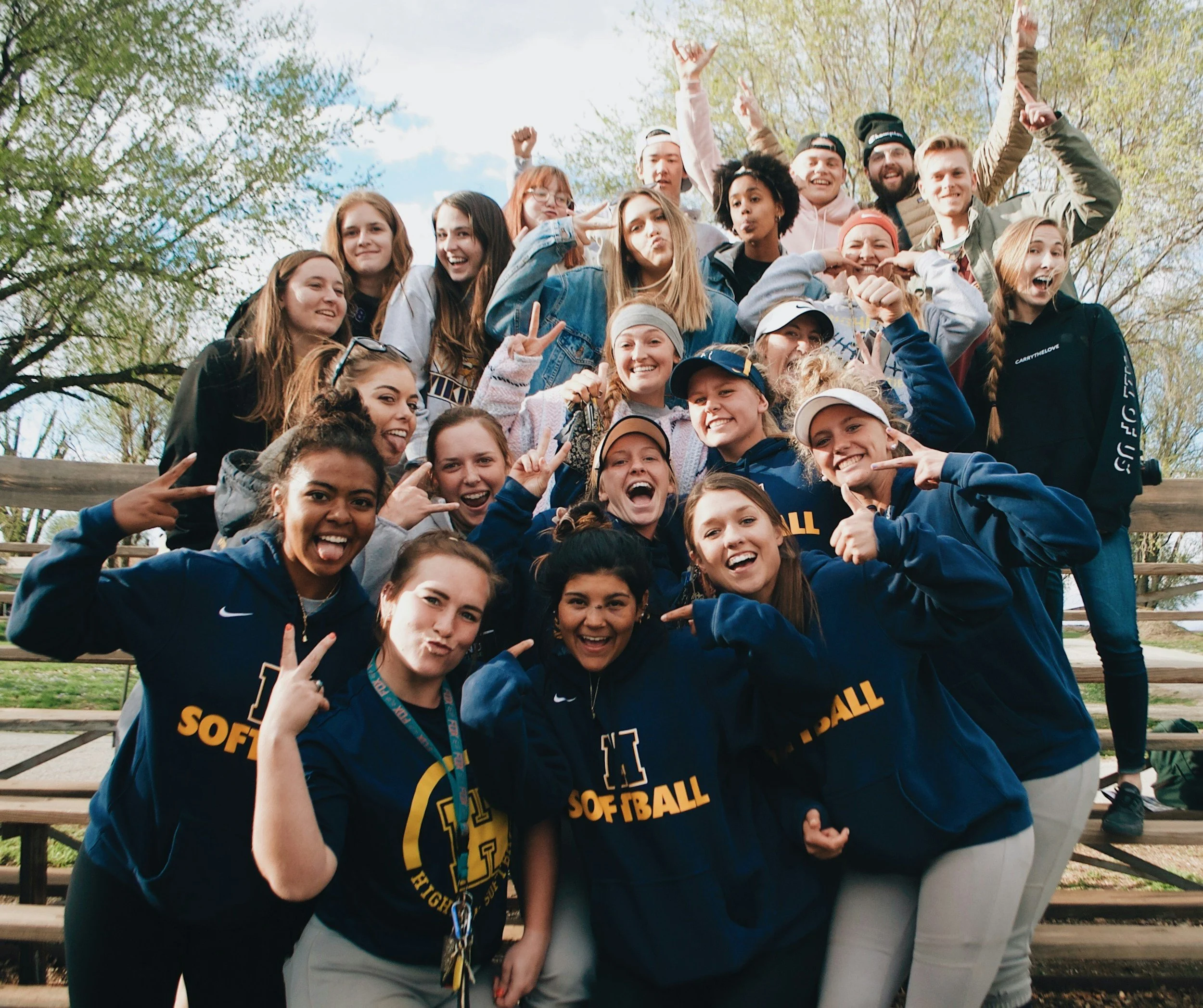Group of smiling young people posing outdoors, wearing blue and yellow softball team shirts, making peace signs and various hand gestures, with trees and a cloudy sky in the background.