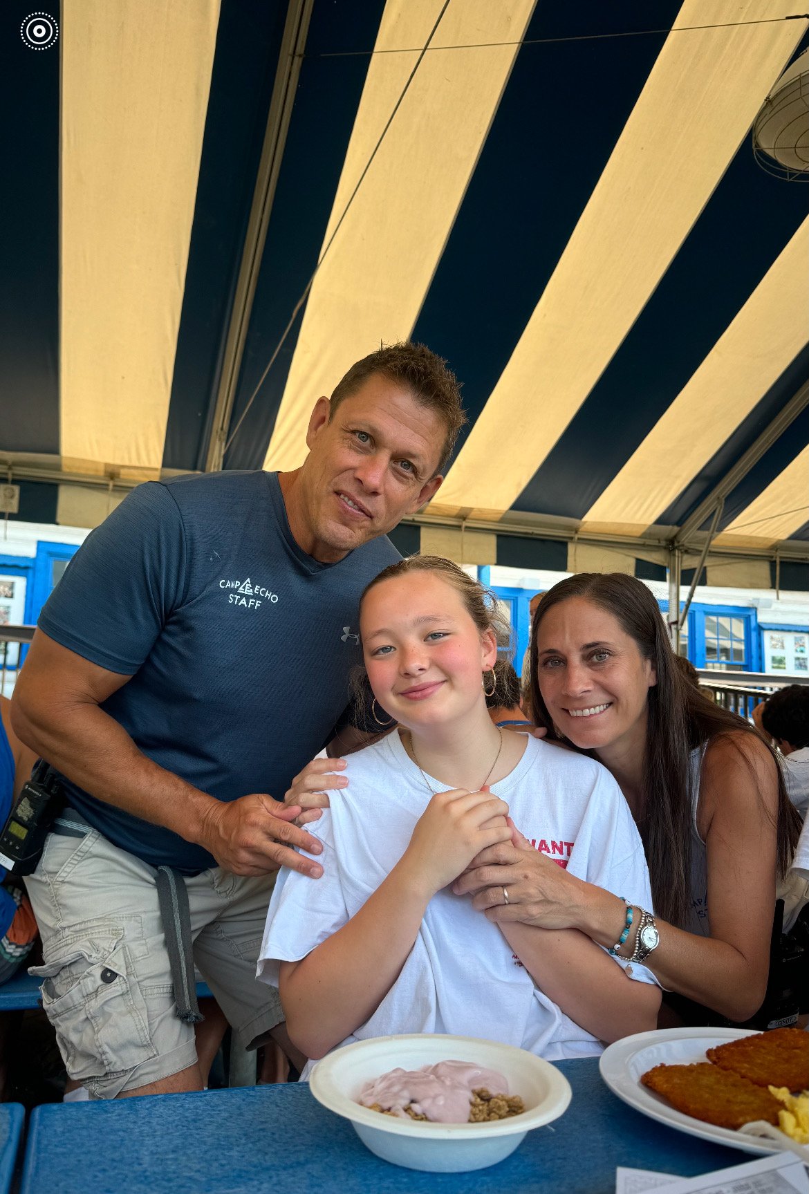 A man, a young girl, and a woman sit together at a table under a large striped yellow and blue tent. The man and woman are smiling, while the girl is looking at the camera. There are plates of food on the table in front of them.