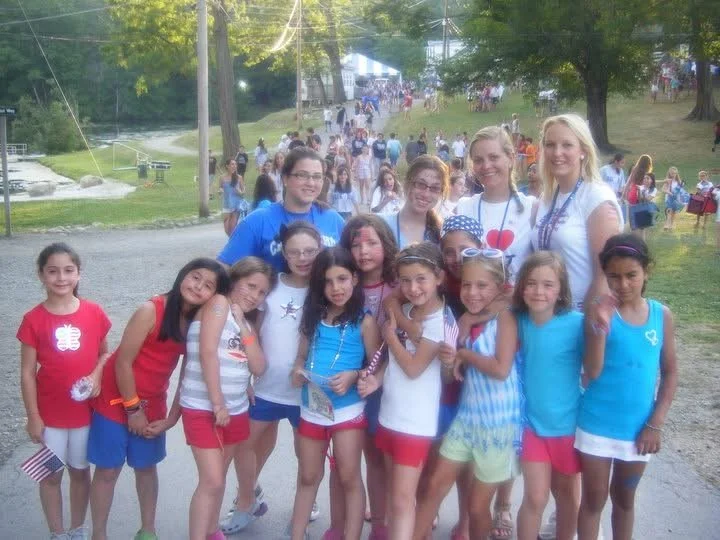 A group of young girls and women gathered outdoors at a summer event with many more people in the background. The girls are wearing colorful summer outfits, and some are holding small American flags.