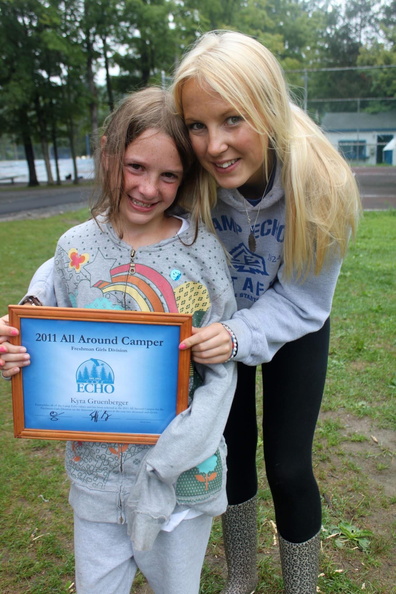 A young girl with Down syndrome holding a certificate, standing beside an older girl with long blonde hair, both smiling outdoors on a grassy area with trees in the background.