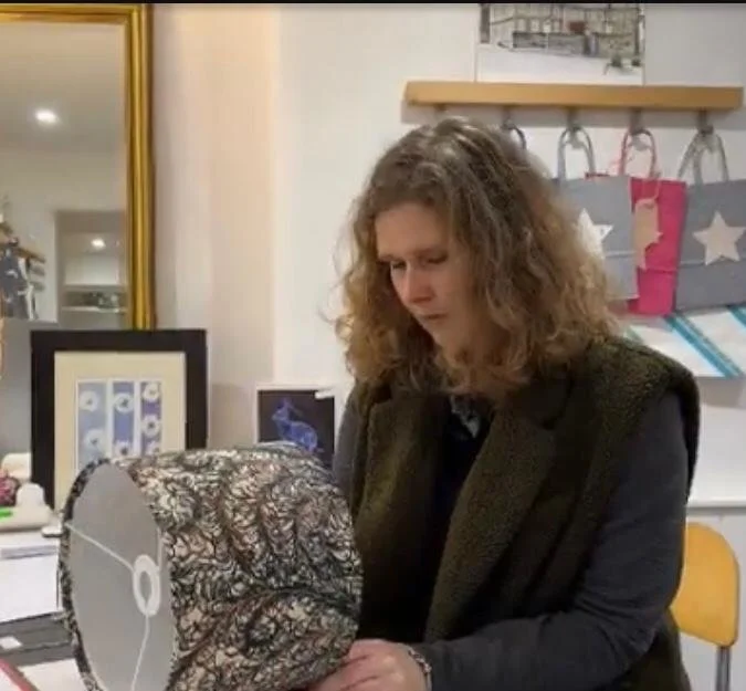 A woman with curly hair looking at a spherical object covered in black and white patterned fabric on a table in a shop or studio.