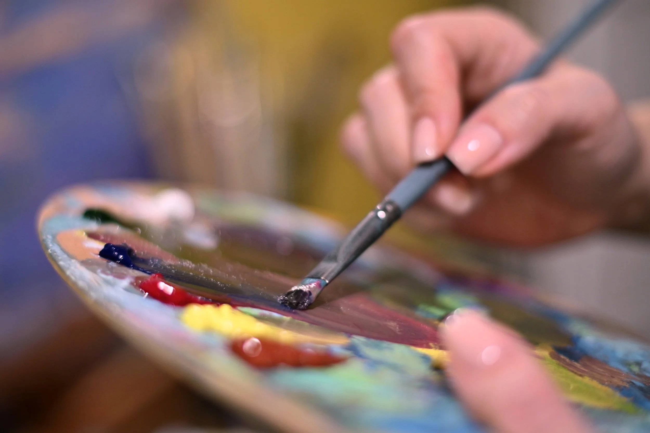 Close-up of a hand holding a paintbrush being dipped into a colorful paint palette.