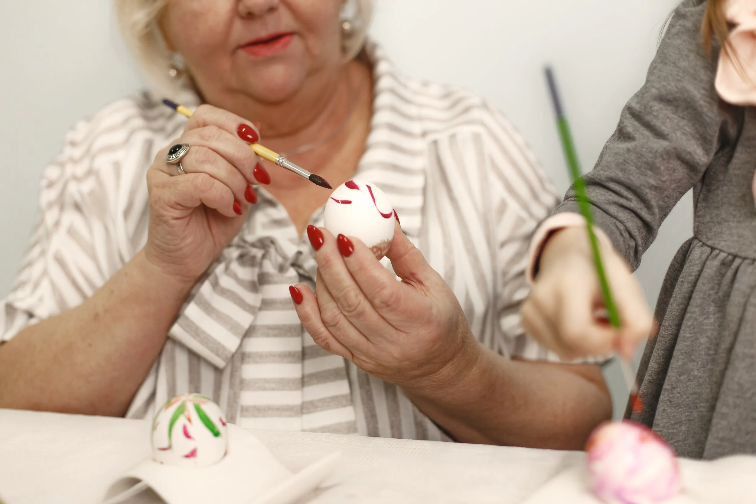 An elderly woman with red nail polish is painting red patterns on an egg, with a young girl assisting. The woman is wearing a striped blouse and has short, light-colored hair. There are decorated eggs on the table.