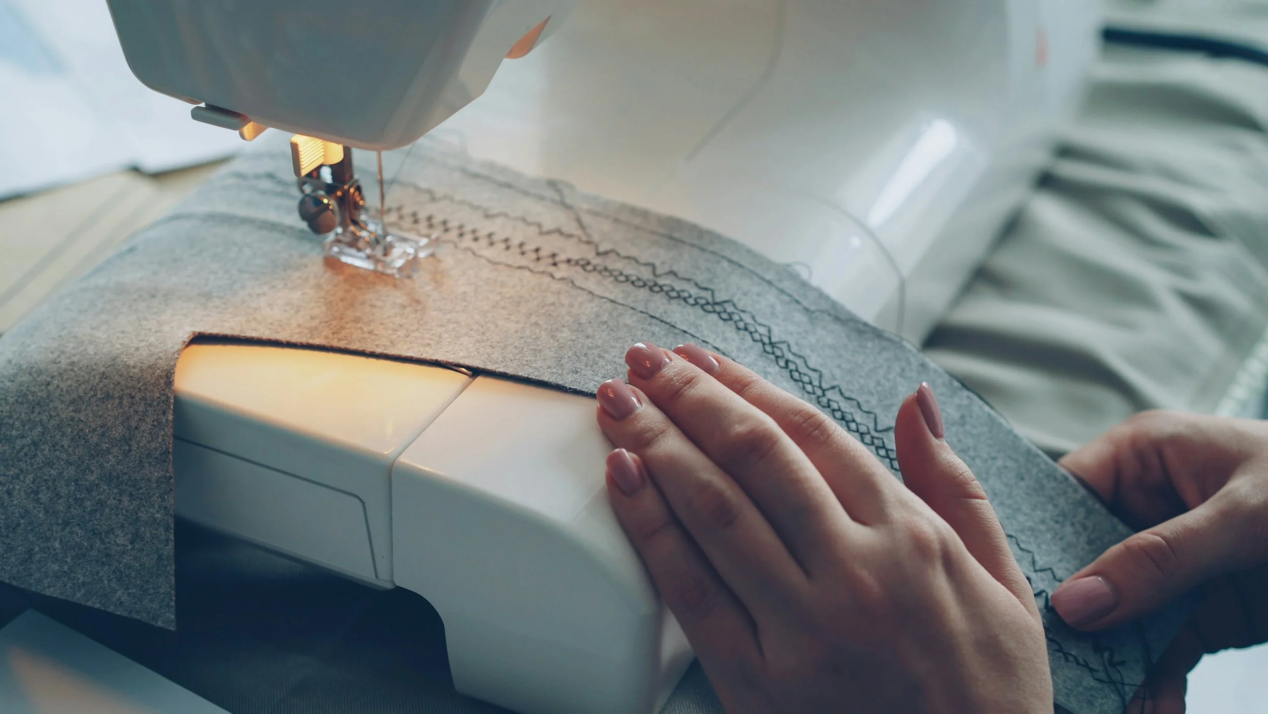 Close-up of hands guiding fabric through sewing machine, stitching decorative patterns on a piece of gray fabric.