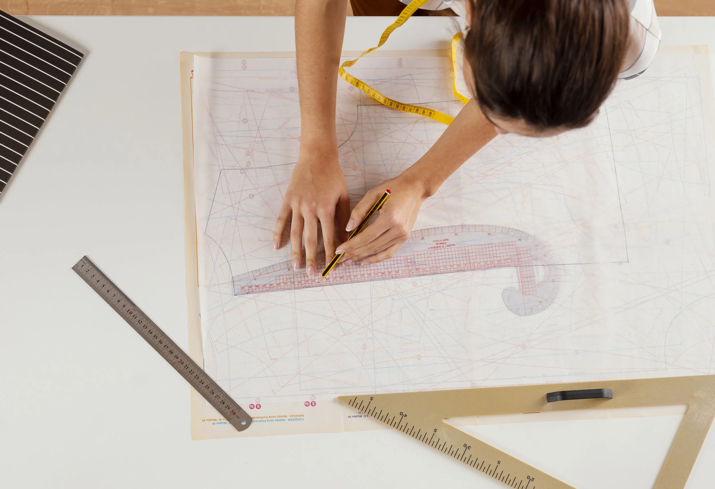 Top-down view of a person working on a sewing pattern, using a ruler, a pencil, and a measuring tape on a large template with drafting lines and markings, with additional rulers nearby on a white work surface.