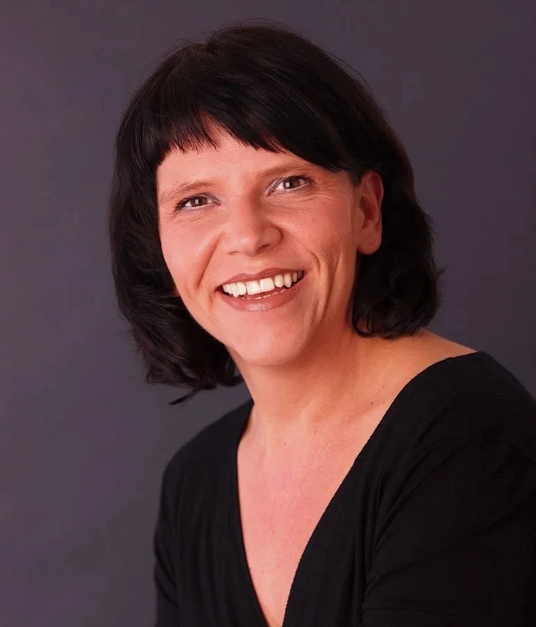 Porträt einer lachenden Frau mit schwarzen, kurzen Haaren vor grauem Hintergrund.