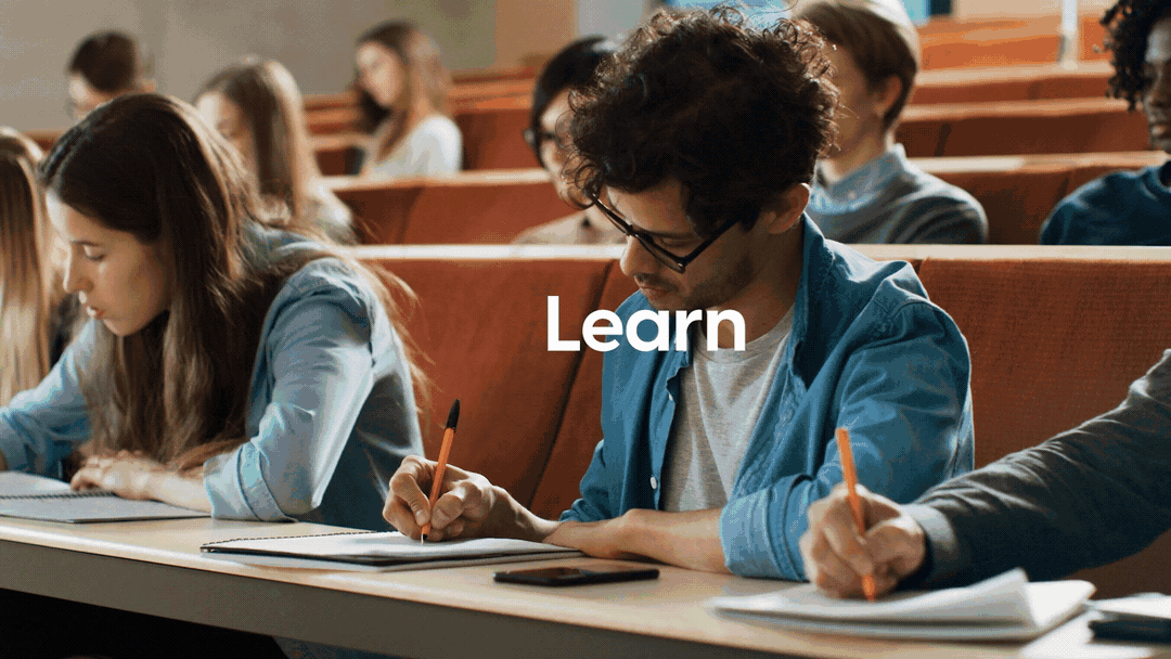 Students taking notes in a classroom or lecture hall with a focus on a young man with glasses and curly hair, wearing a blue jacket, and a young woman with long brown hair.