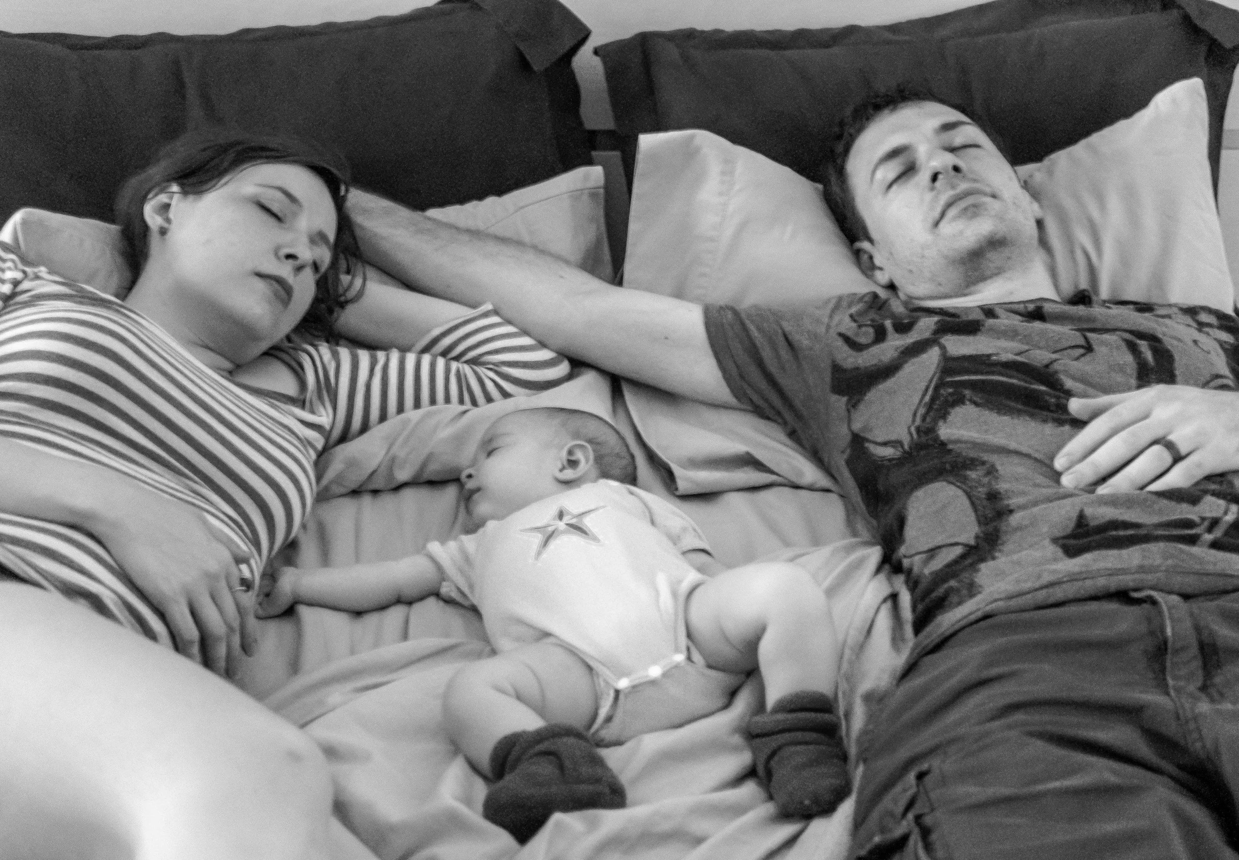 A mother and father asleep on a bed with their baby sleeping between them
