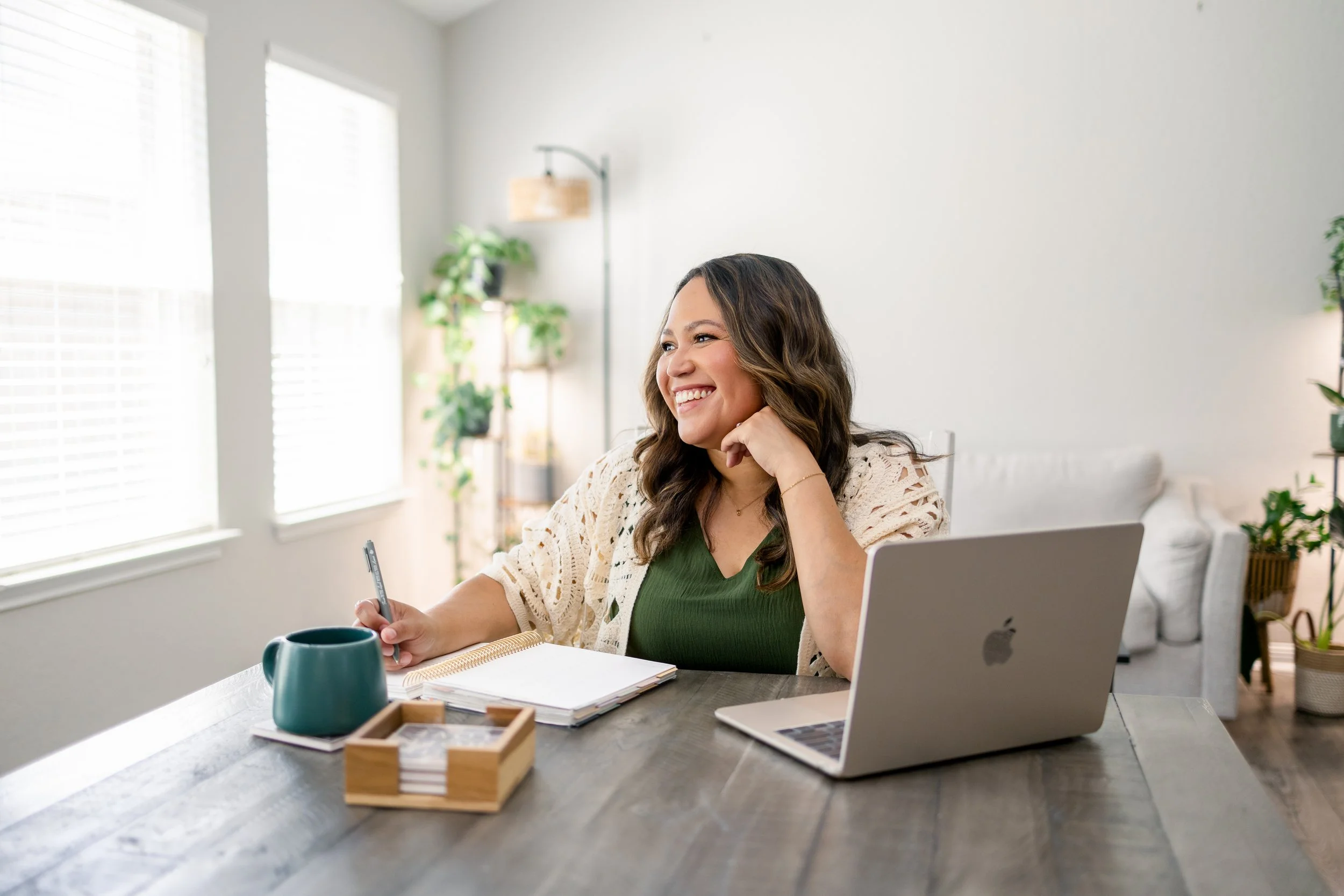 A woman with a bun hairstyle sitting on a chair, holding a young boy, in front of a gray brick wall, using a laptop placed on a white table, with her right hand pointing at the laptop's trackpad.