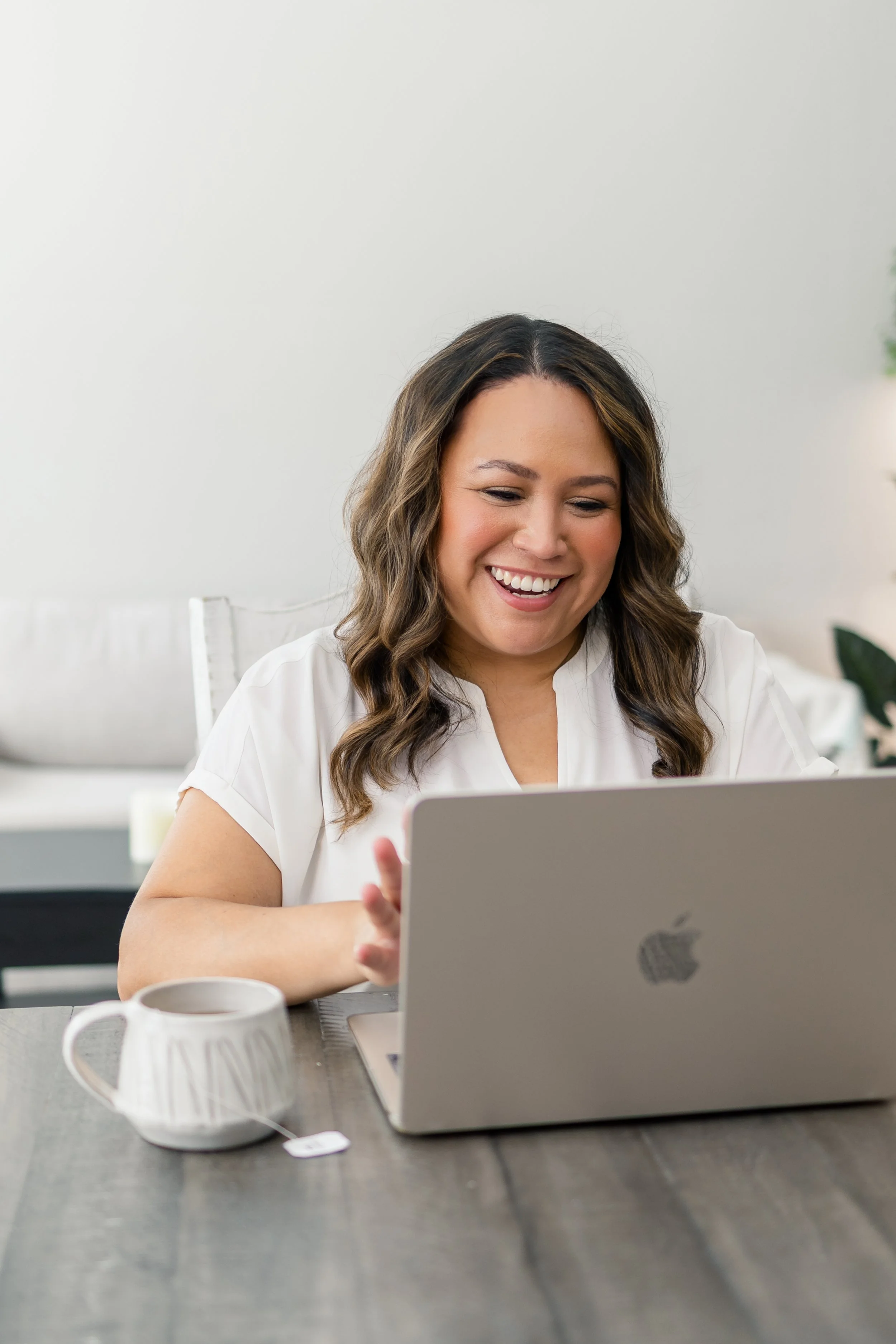 A woman with glasses and earrings sitting at a table with a laptop, holding a cup, smiling, and waving. On the table are a plate with bread, a glass jar of red liquid, and a glass pitcher. In the background, part of a kitchen is visible with cabinets and decorative items.