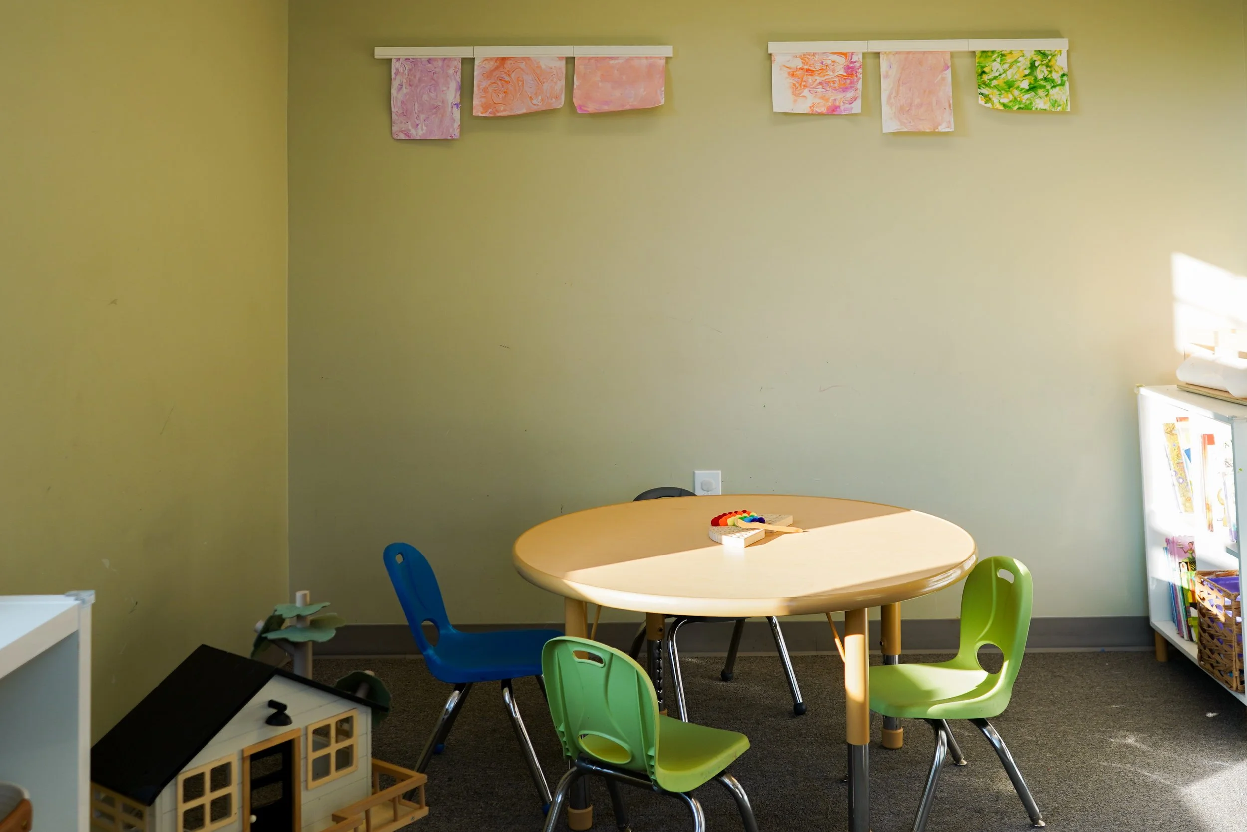 Children's playroom with a round table, colorful chairs, toys, and children's artwork hanging on the wall.