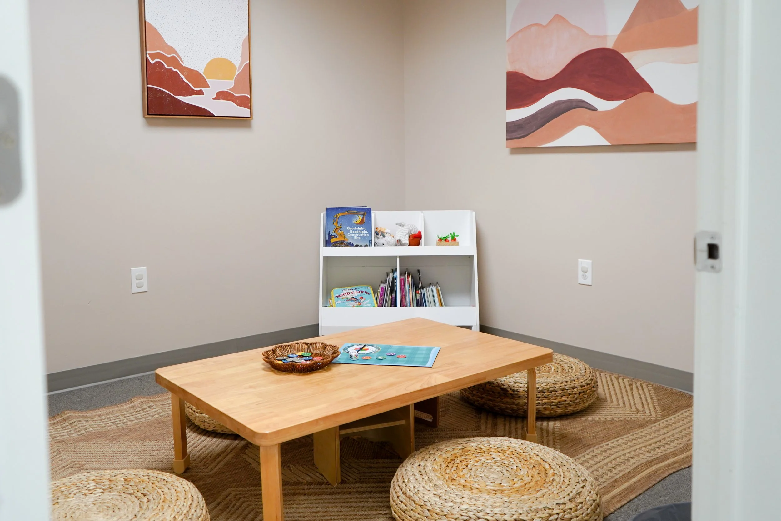 A cozy children's reading nook with a low wooden table, woven round cushions, a small white bookshelf filled with children's books and toys, two abstract landscape paintings on the walls, and a woven rug on the floor.