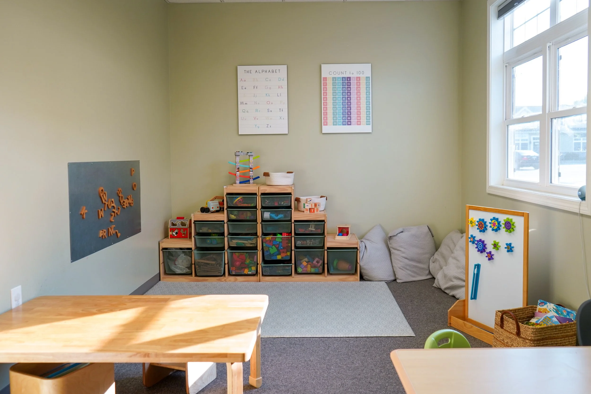 A daytime classroom corner with educational posters on the wall, toy storage bins, bean bags, a whiteboard with magnetic decoration, and a window with natural light.