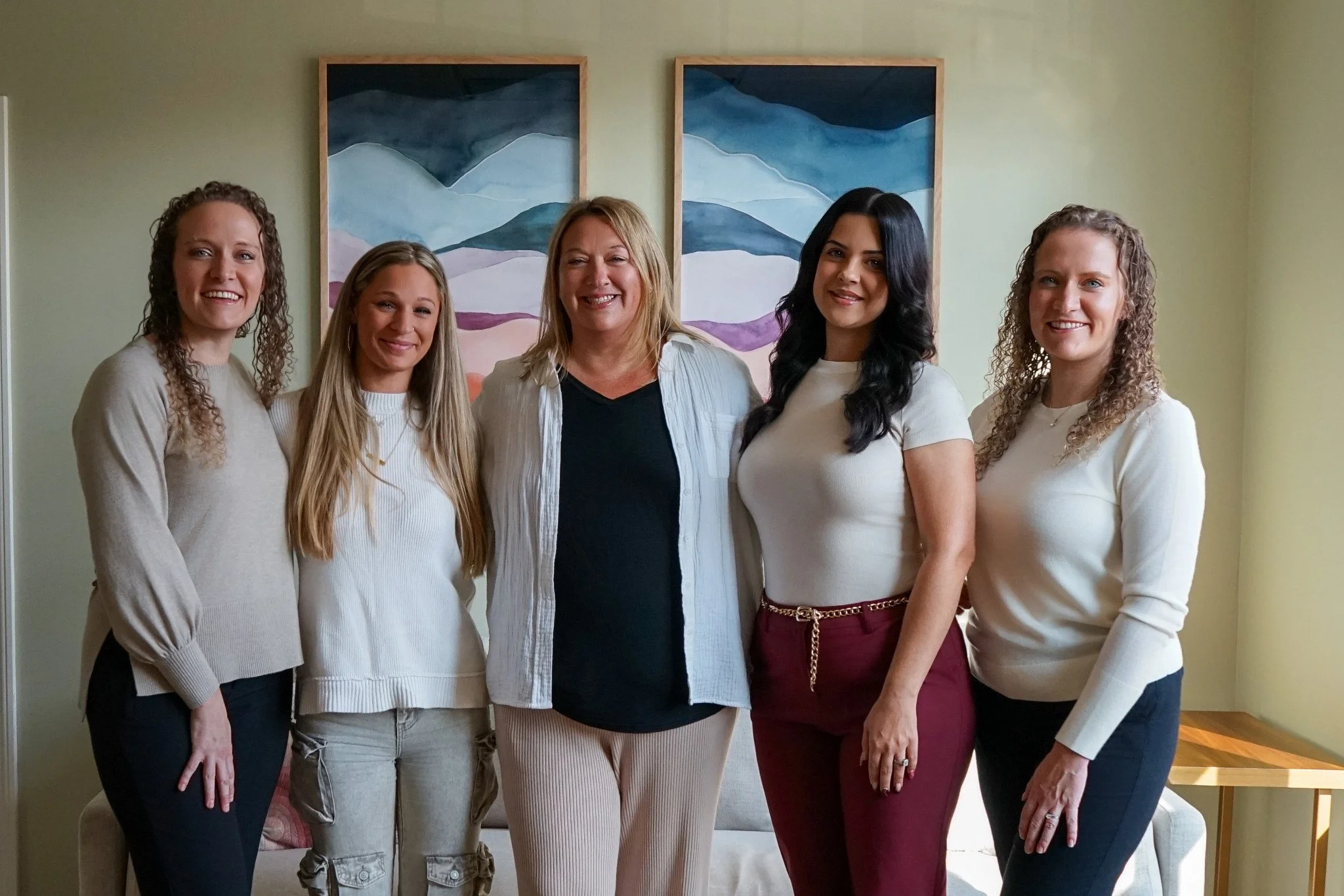 Group of five women standing together indoors, smiling at the camera, with two mountain landscape paintings hanging on the wall in the background.