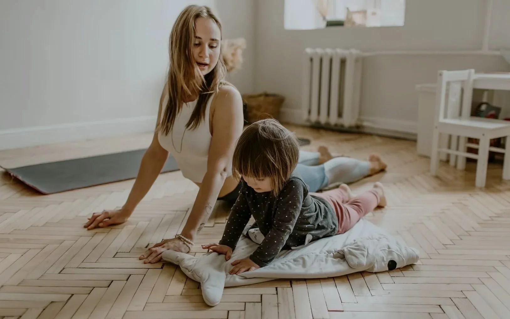 A woman and a young girl doing yoga on a wooden floor in a bright room.
