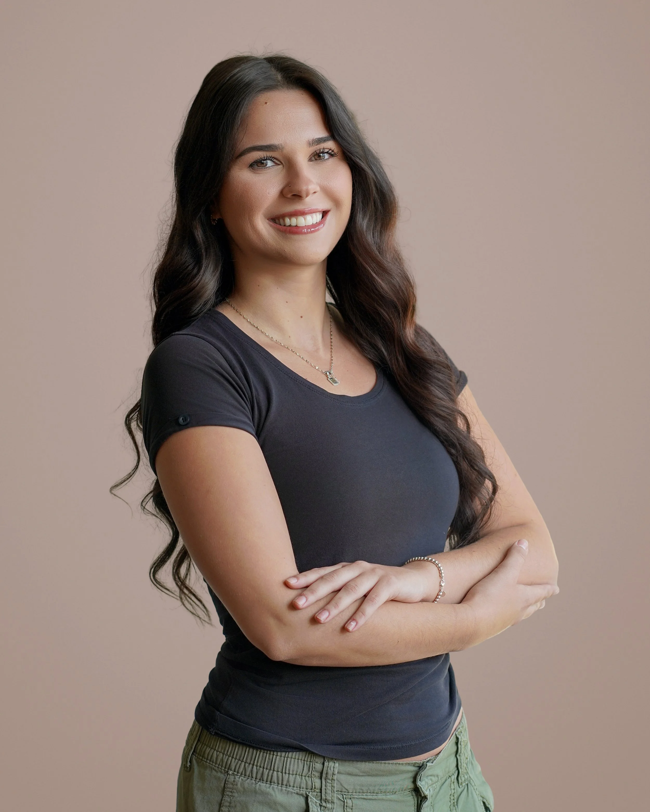 A young woman with long, wavy dark hair smiling, standing with her arms crossed against a neutral background.