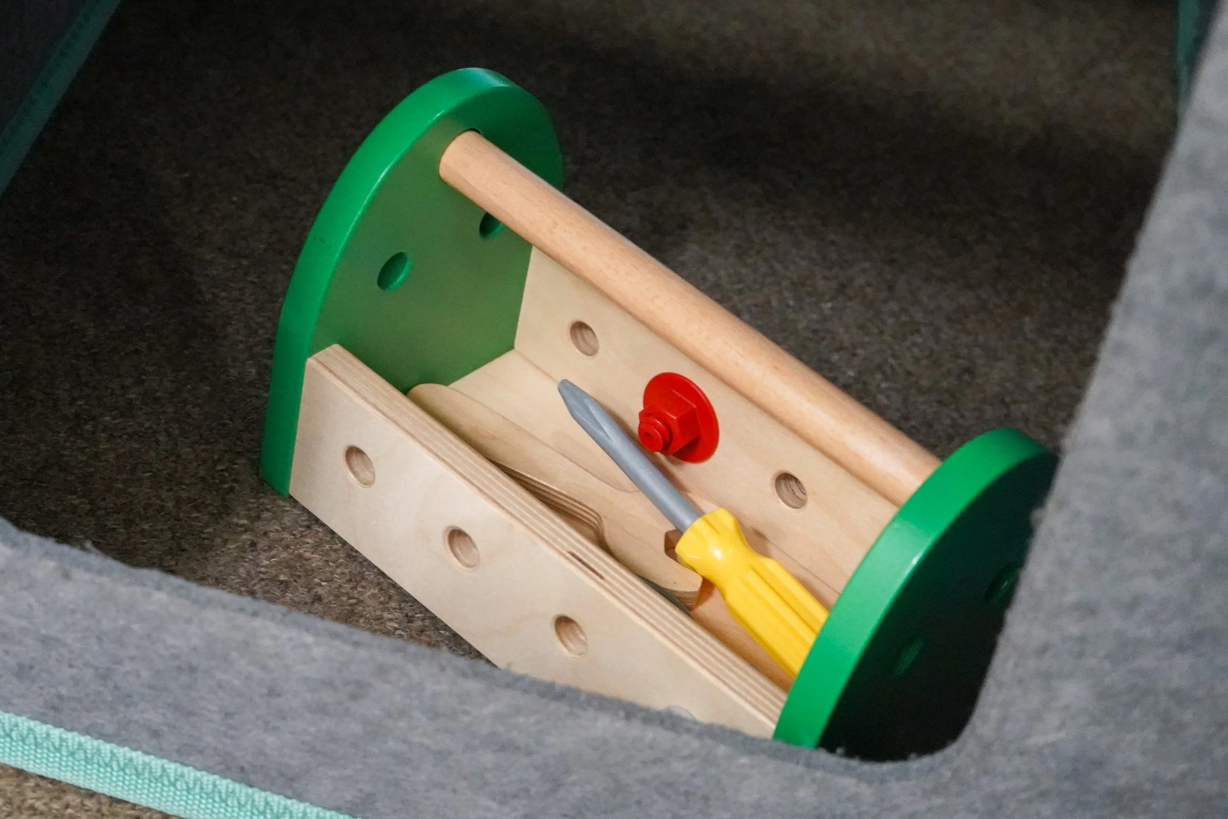 Unfinished wooden toy with green wheels, a yellow screwdriver, and a red bolt, placed inside a gray fabric-lined box.
