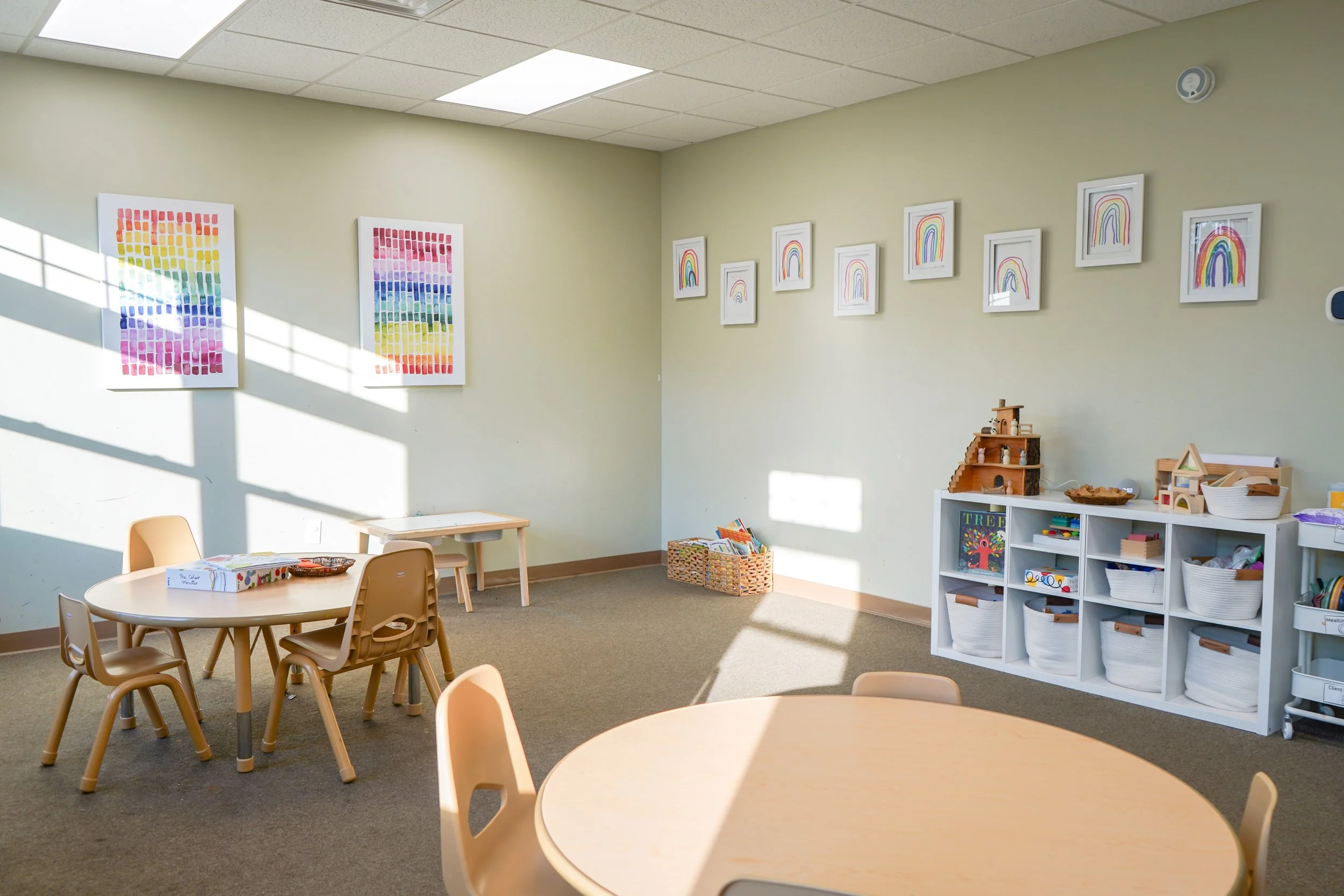 A brightly-lit preschool classroom with colorful wall art, light green walls, and wooden furniture, including small tables and chairs, with toys and baskets on white shelves.