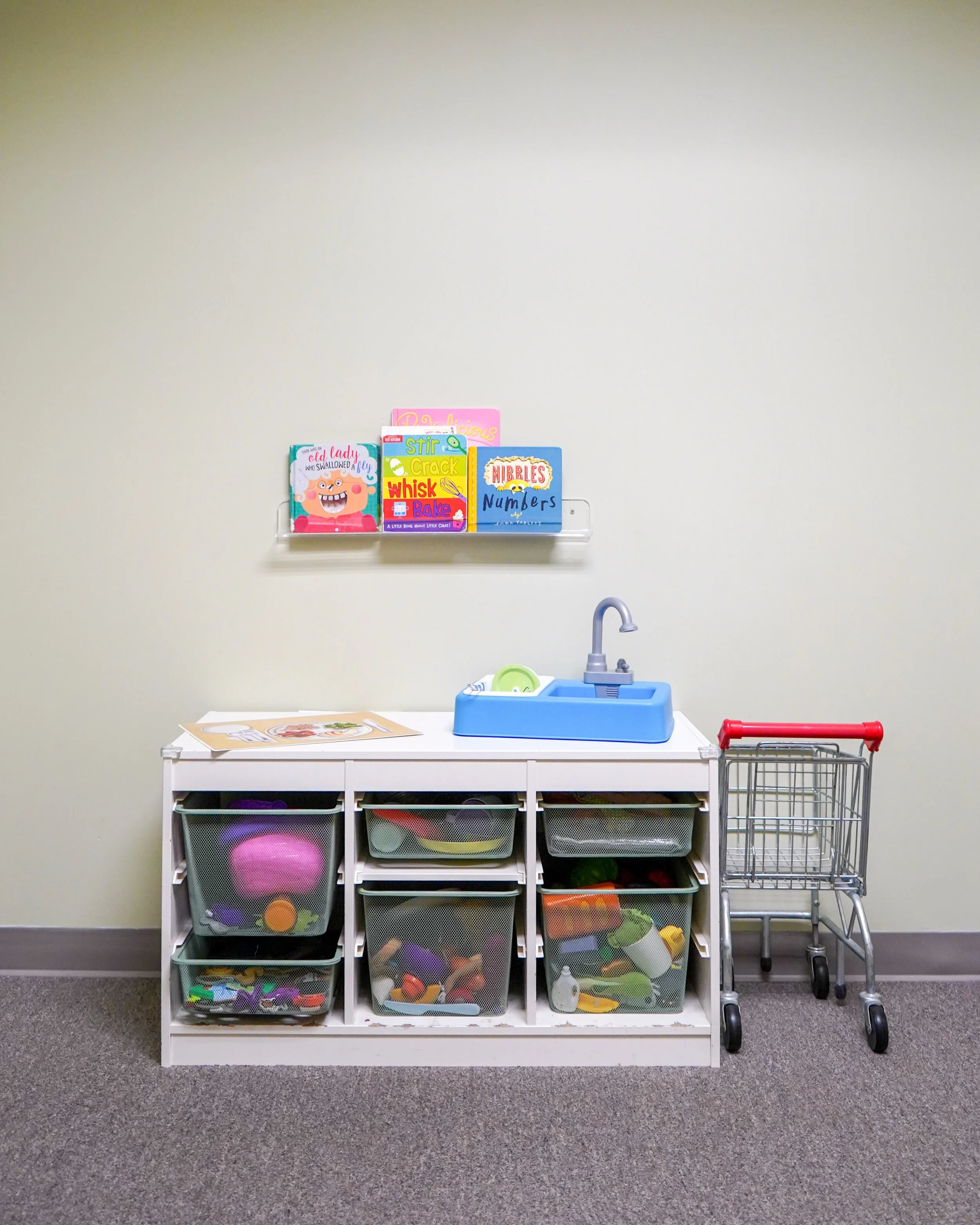 A small white shelf with mesh baskets filled with colorful children's toys, next to a small play sink with a blue basin and purple faucet, set against a plain beige wall with children's books displayed on a wall shelf above.