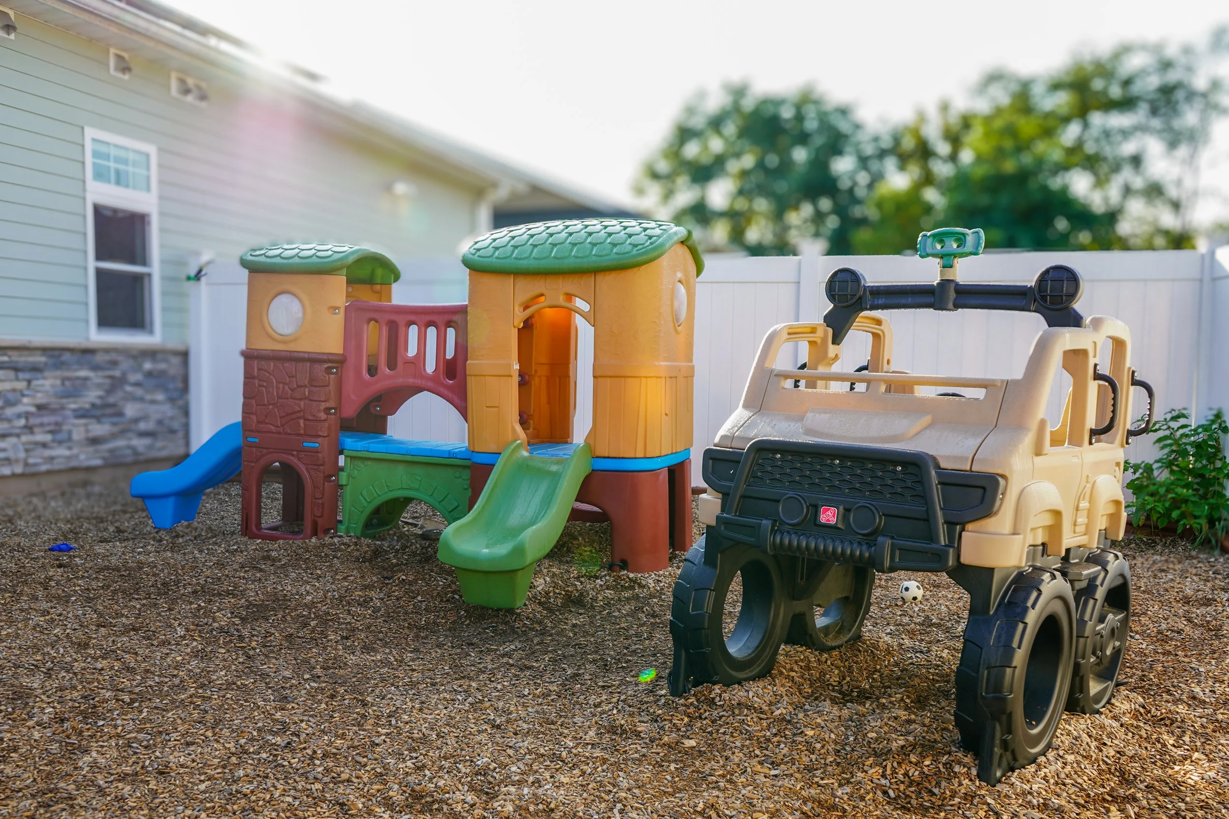 Children's outdoor play area with a colorful plastic playhouse with slides and a ride-on toy vehicle on a gravel surface, surrounded by a white fence and house with siding in the background.