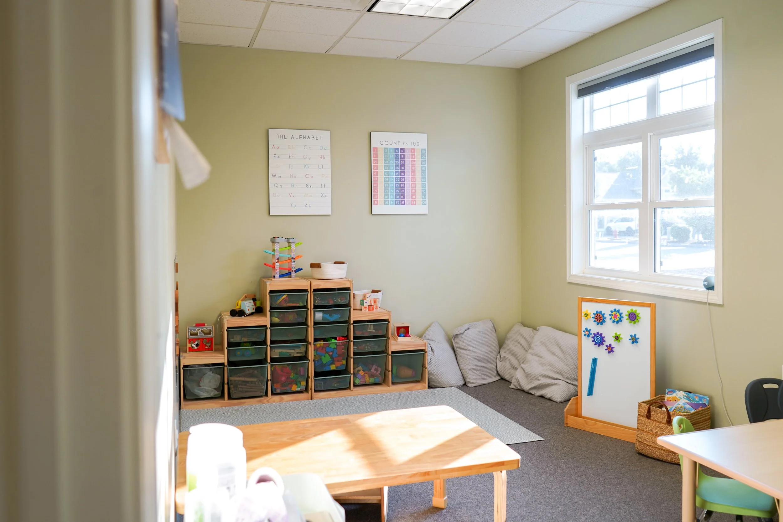 A near-empty classroom with light green walls, a large window, posters on the wall, a wooden shelf filled with plastic bins of toys and educational materials, a small whiteboard with magnetic decorations, and a row of cushioned pillows on the floor.