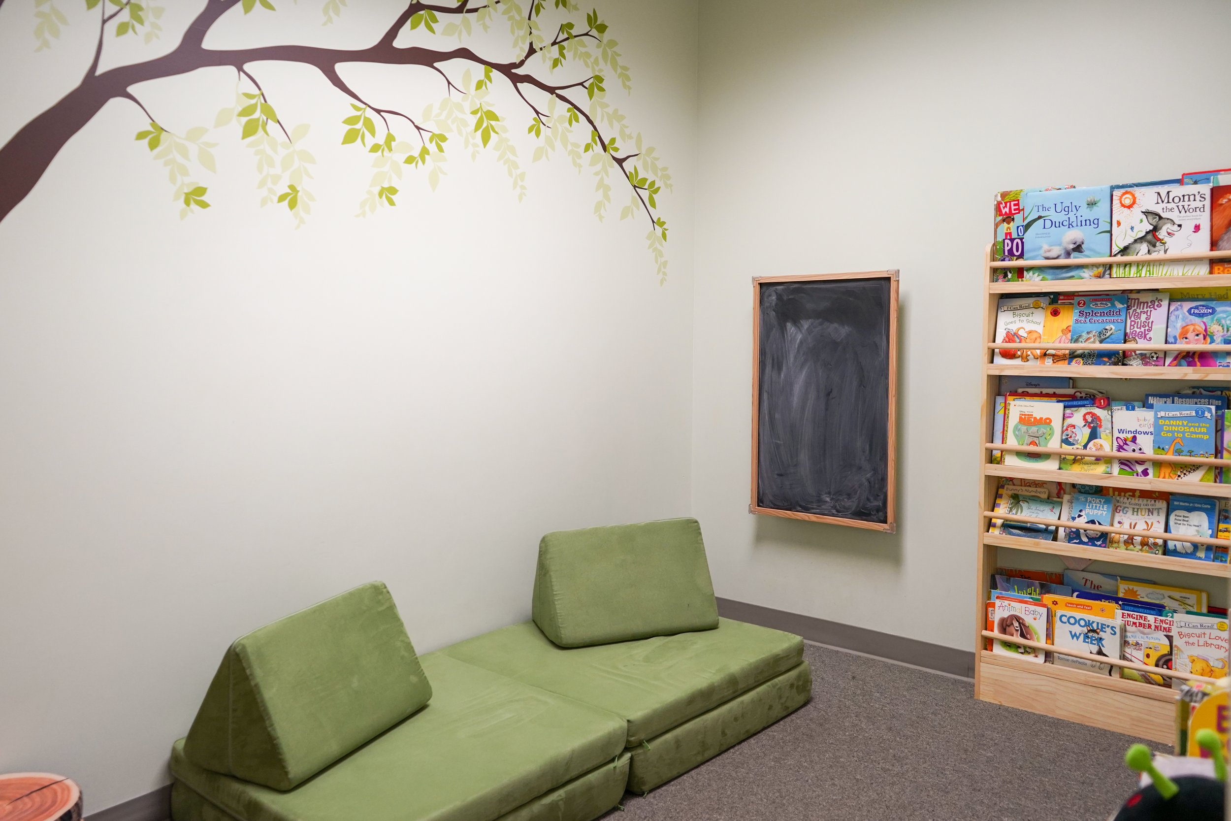 A corner of a children's reading room with a green sofa that has three cushions, a small chalkboard on the wall, and a wooden bookshelf filled with colorful children's books. The wall features a large painted tree branch with green leaves.