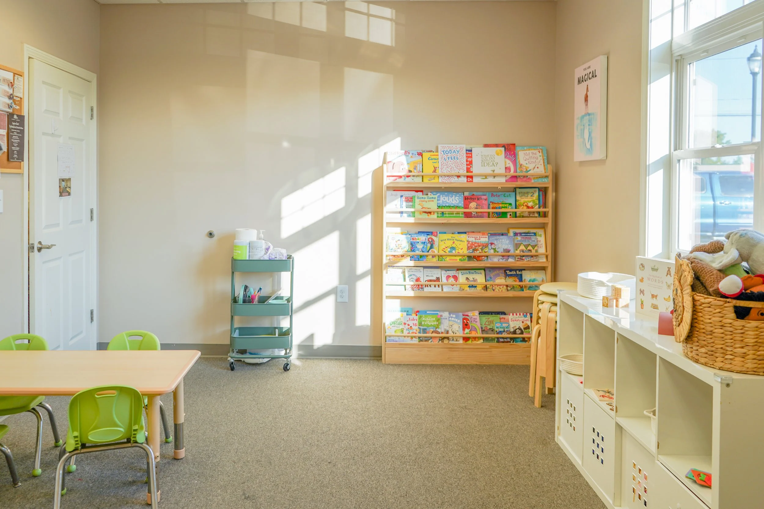 A bright children's reading room with a small table and green chairs, a bookshelf filled with children's books, a window with sunlight, and a white storage unit with baskets and toys.