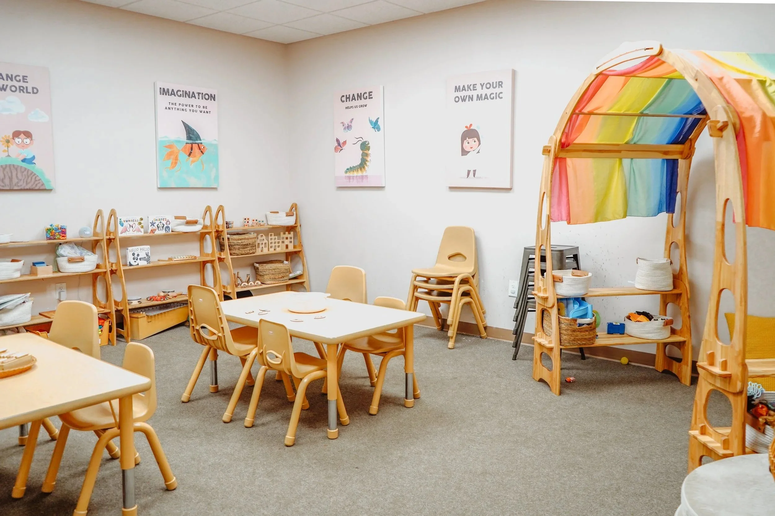 An empty classroom with small tables and chairs, wooden shelves filled with baskets of toys and supplies, and colorful posters on the white walls.
