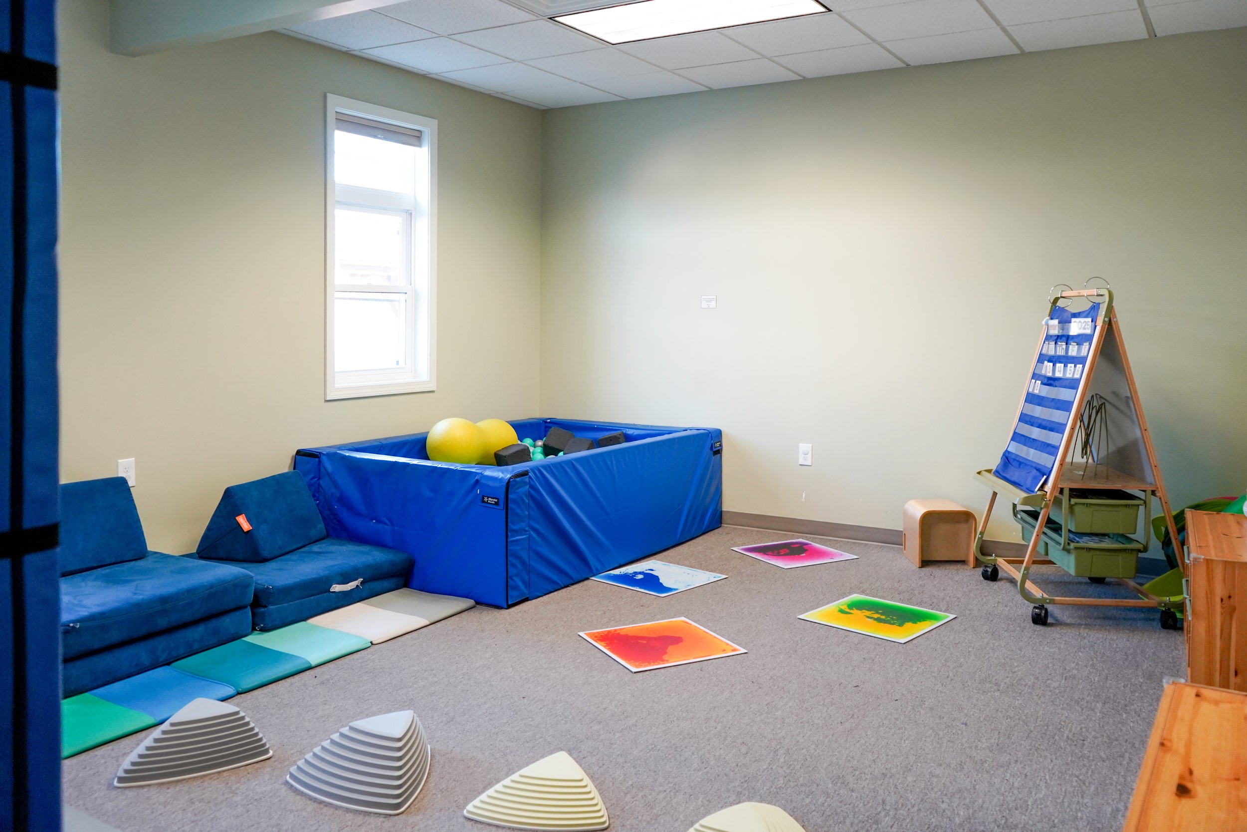 Empty children's play room with blue foam mats, colored skill tiles, a small blue ball pit with yellow balls and black and green toys, a blue couch with foam wedges, and a wooden A-frame cart with blue hanging storage.