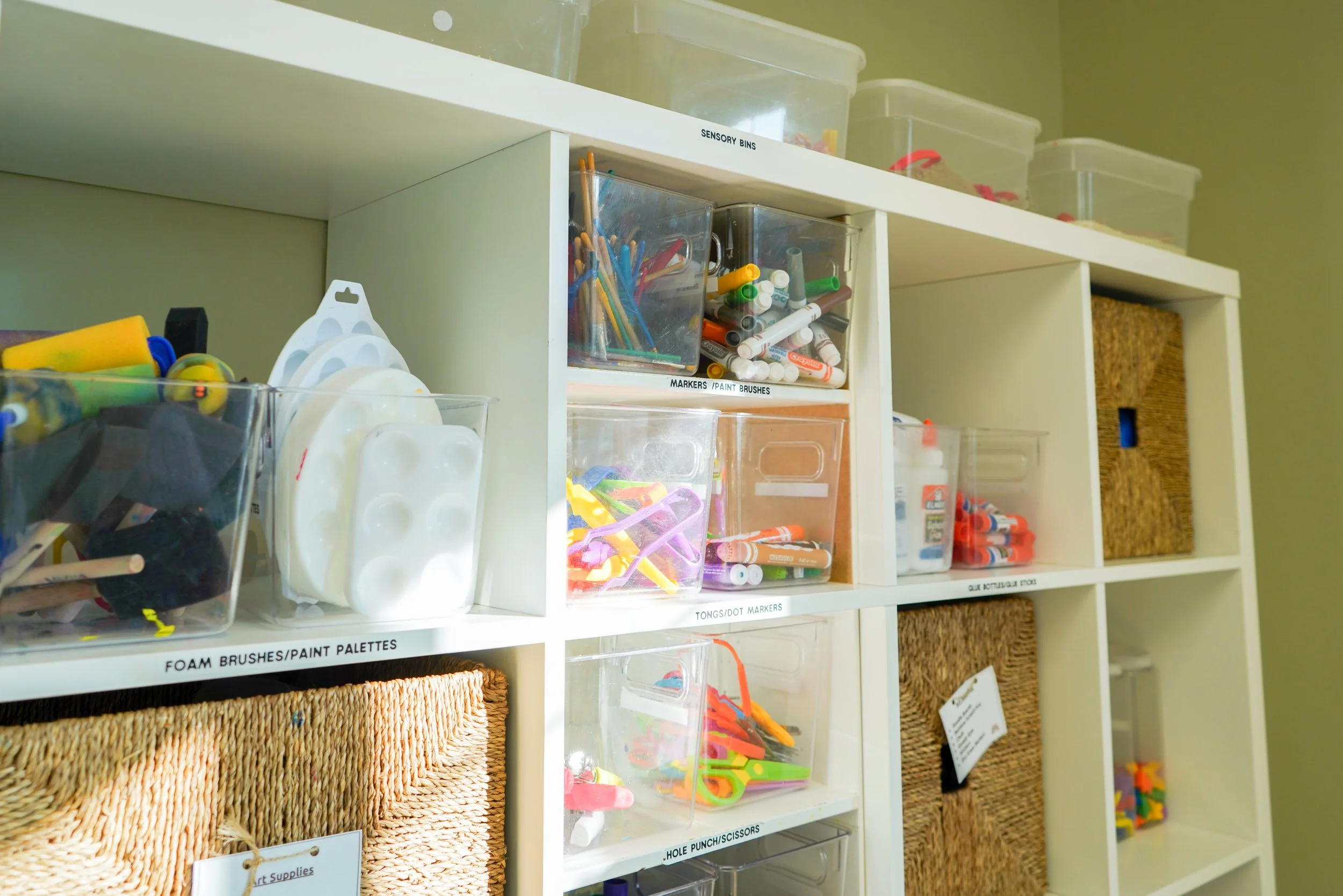 White shelving unit containing various transparent plastic bins filled with art supplies such as paint brushes, markers, glue, and foam brushes, with woven baskets on some compartments.