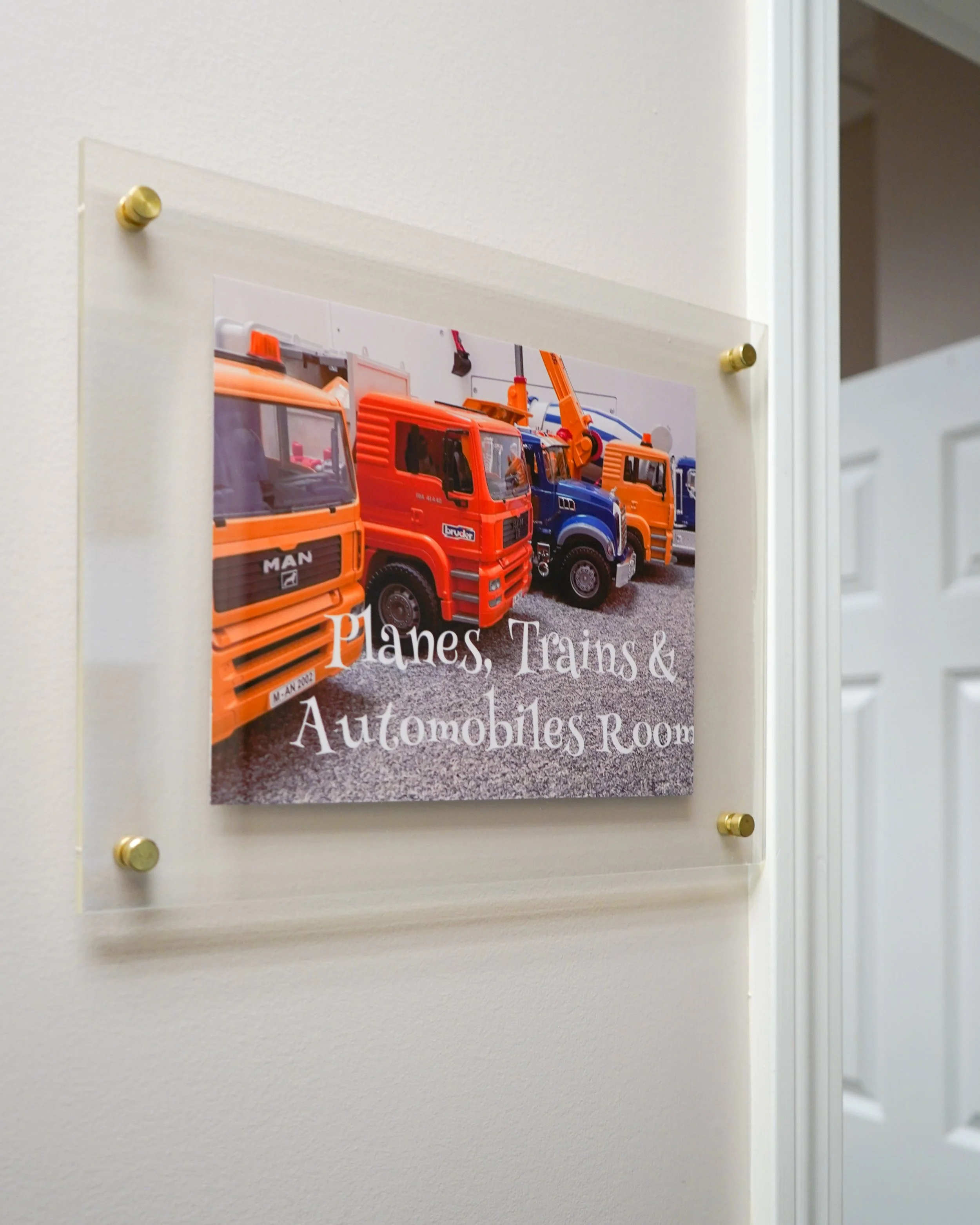 Sign on the wall for planes, trains, and automobiles room with toy vehicles and a white door in the background.