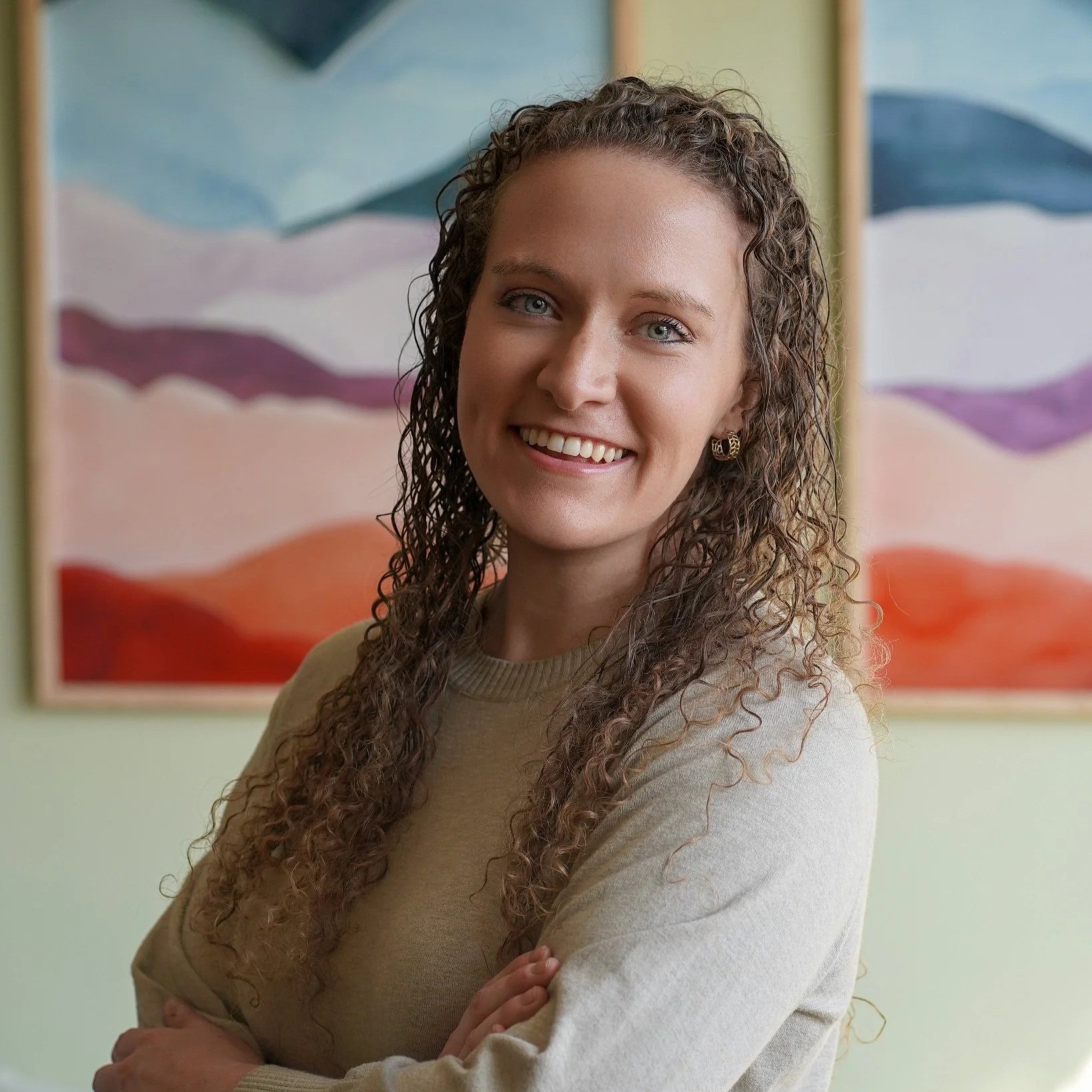 A smiling woman with curly hair and wearing a beige sweater, standing in front of colorful abstract art.