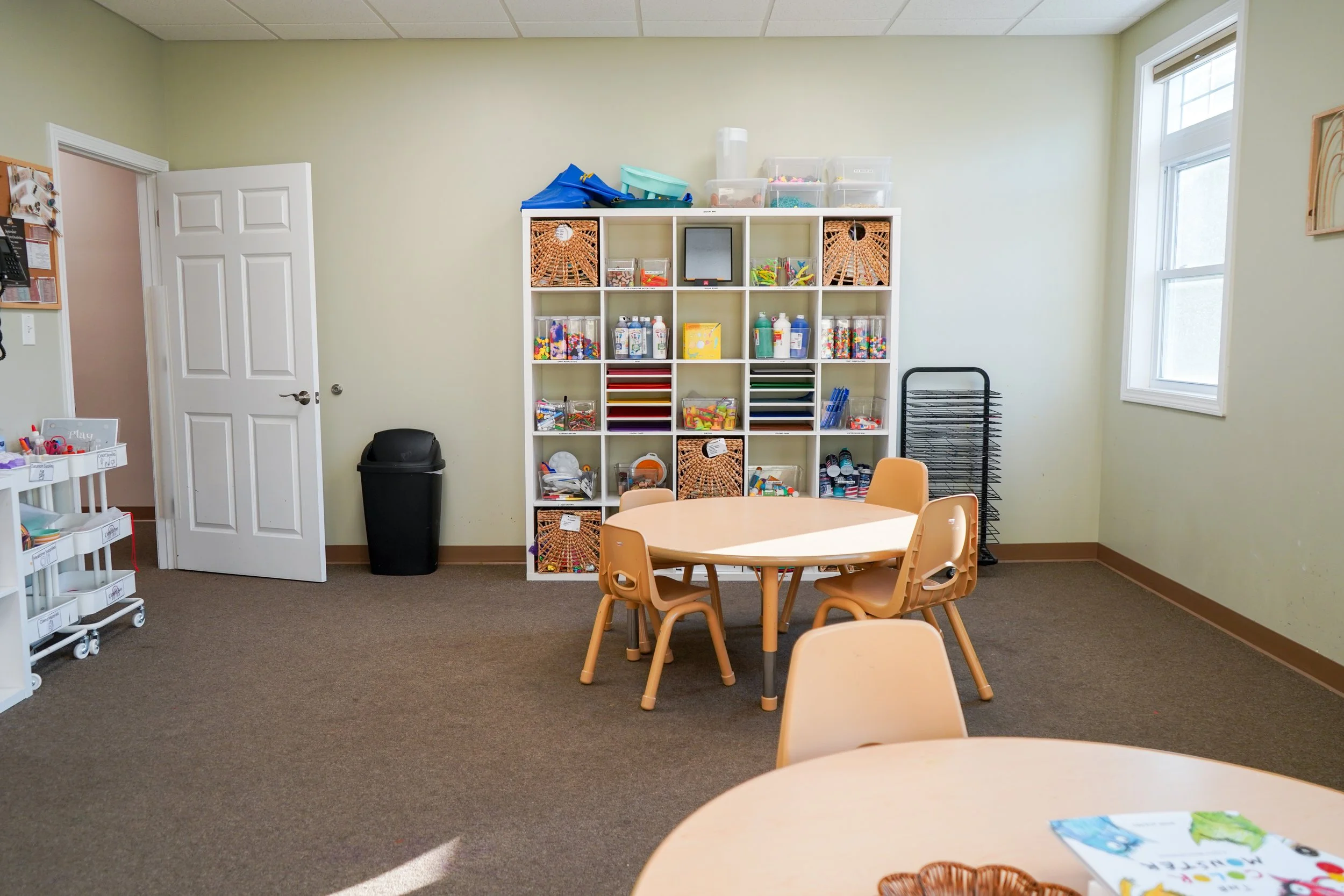 Empty classroom with round tables, plastic chairs, and organized storage shelves filled with art supplies and educational materials.