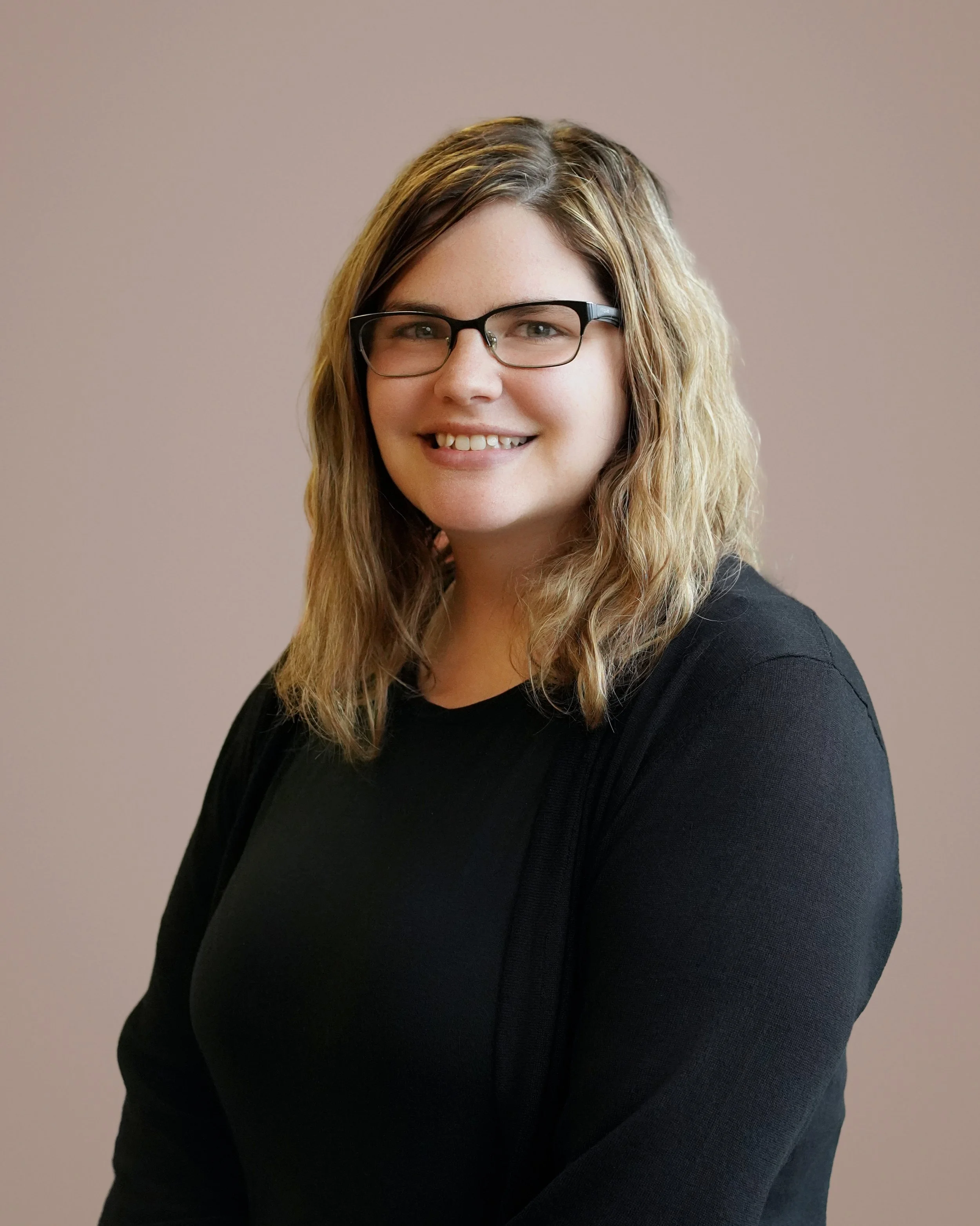 A woman with wavy blonde hair, wearing glasses and a black top, smiling against a plain beige background.