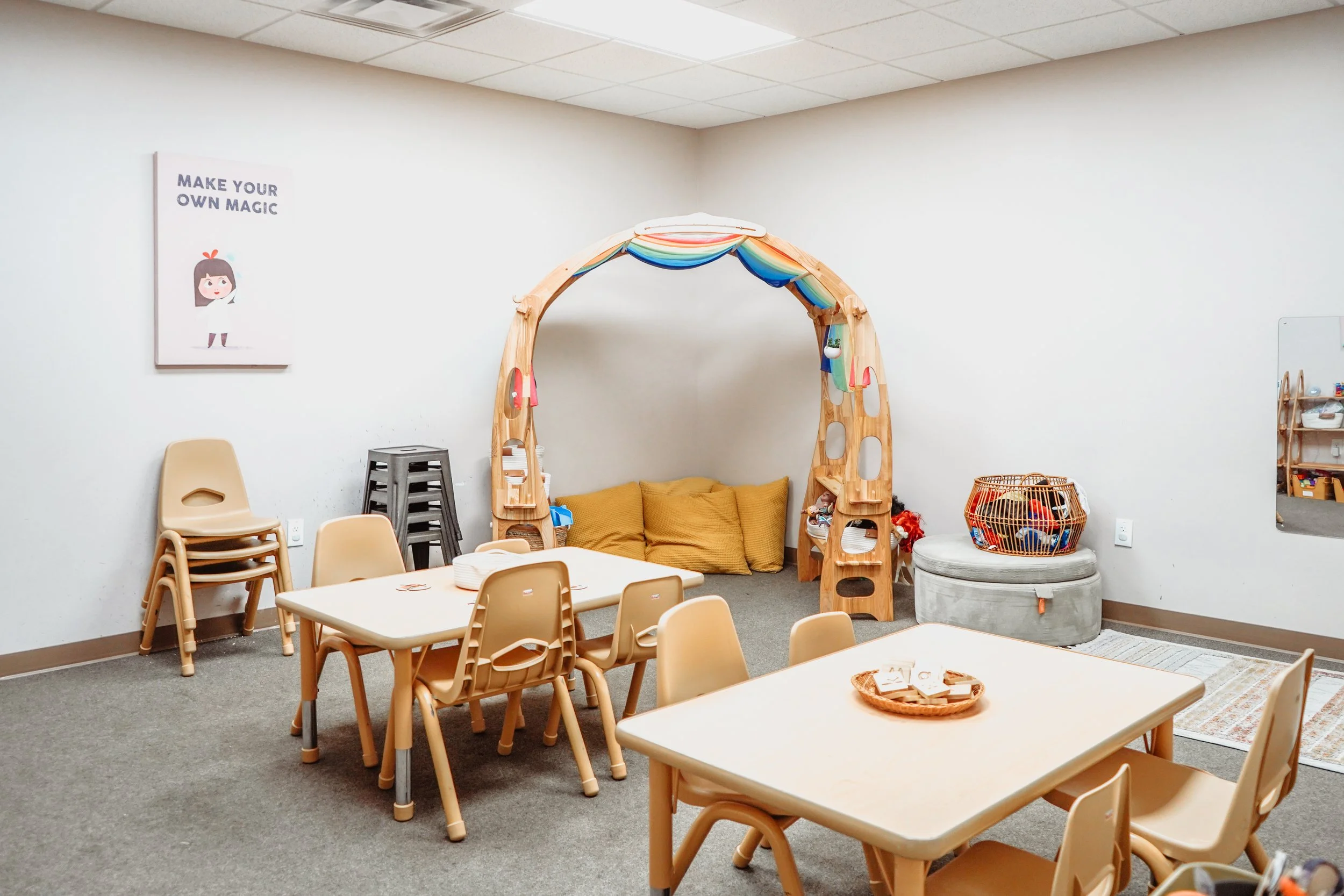 Children's playroom with small tables and chairs, a layered tent with pillows underneath, and a basket of toys.