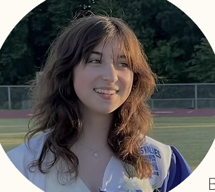 Young woman with brown hair and a white sports uniform standing outdoors on a sports track field, smiling.