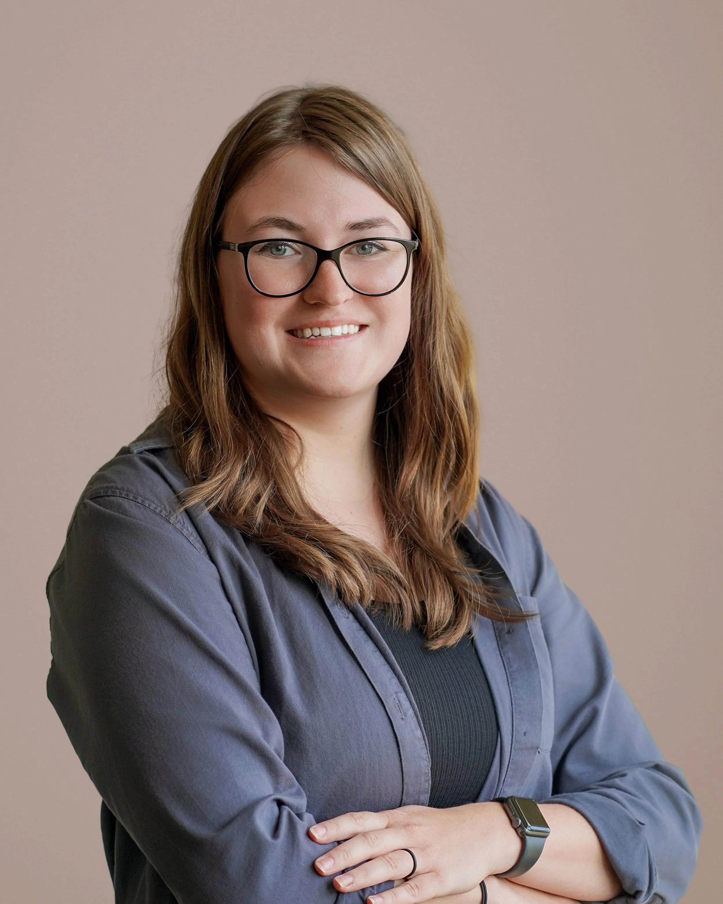 A young woman with glasses and long brown hair, smiling with arms crossed, wearing a dark gray shirt and a smartwatch on her wrist, standing against a neutral beige background.