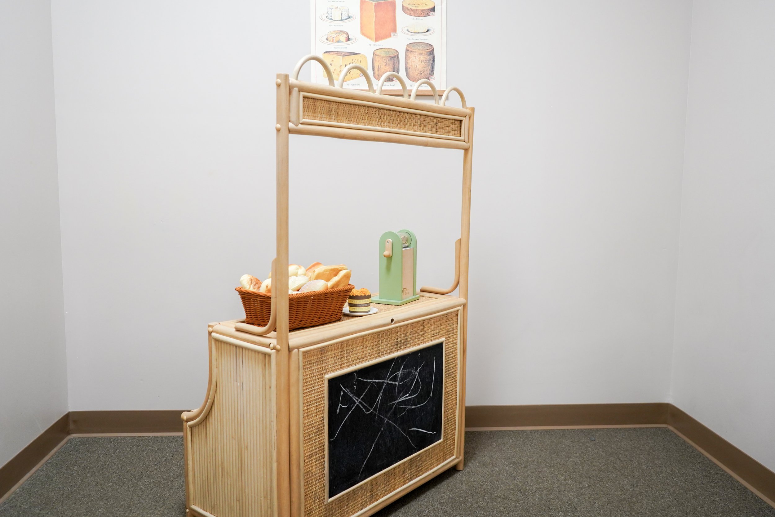 A wooden children's play bakery with a chalkboard front, basket of fake bread, a green toy cash register, a small slice of cake, and a chalkboard sign in a plain room with white walls and gray carpet