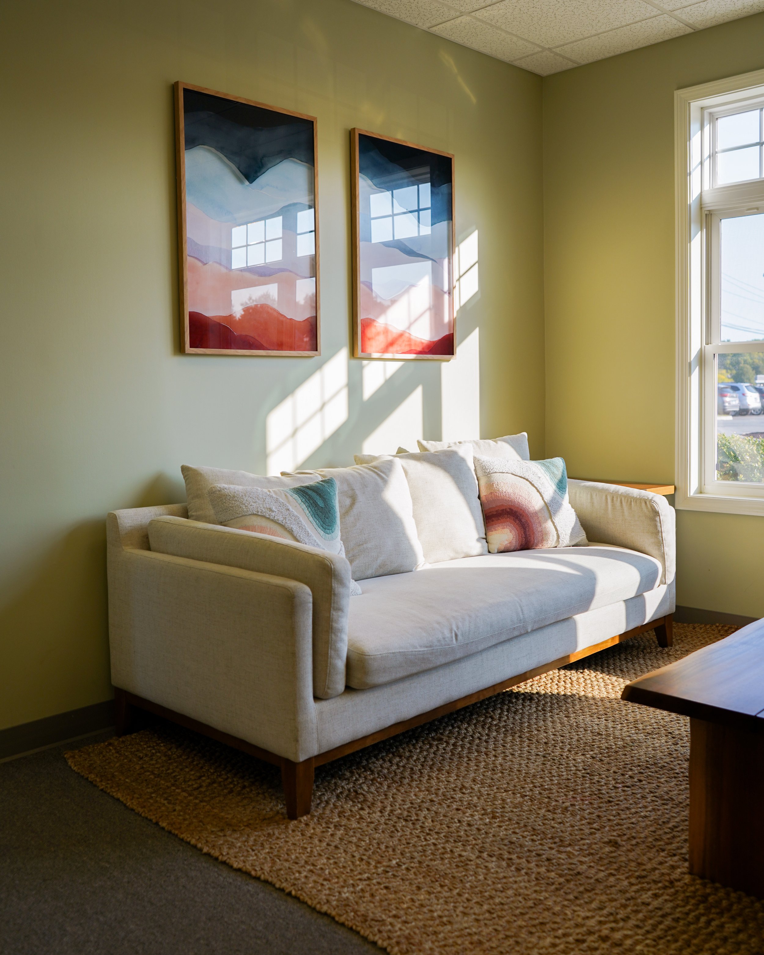 A cozy living room corner with a cream sofa with colorful wave-patterned pillows, a tan woven rug, a wooden coffee table, a light green wall with two abstract mountain landscape paintings, and a window with sunlight streaming in.