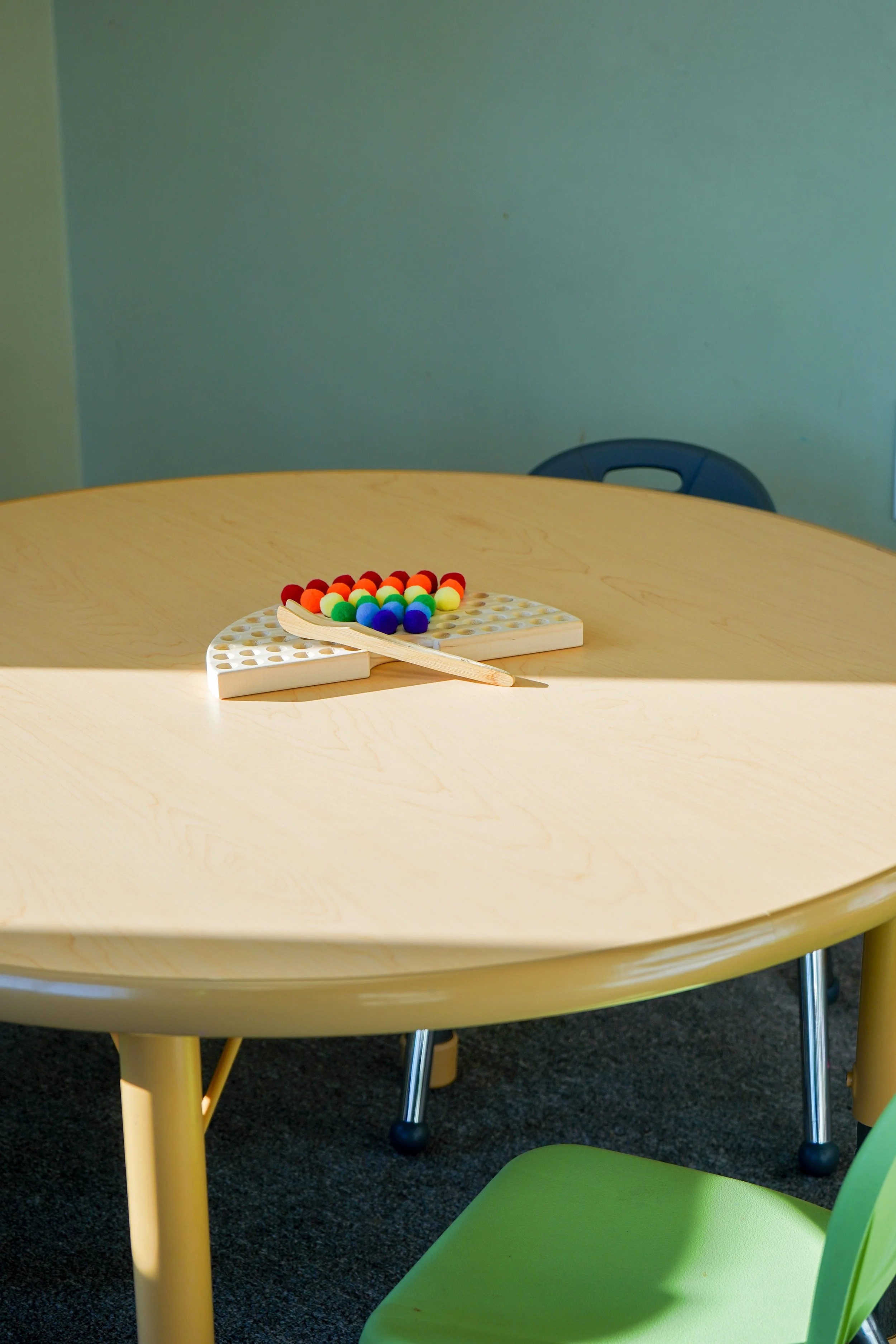 A round table with a game of Montessori rainbow color sorting set, a wooden spatula, and a green chair partially visible in the foreground.