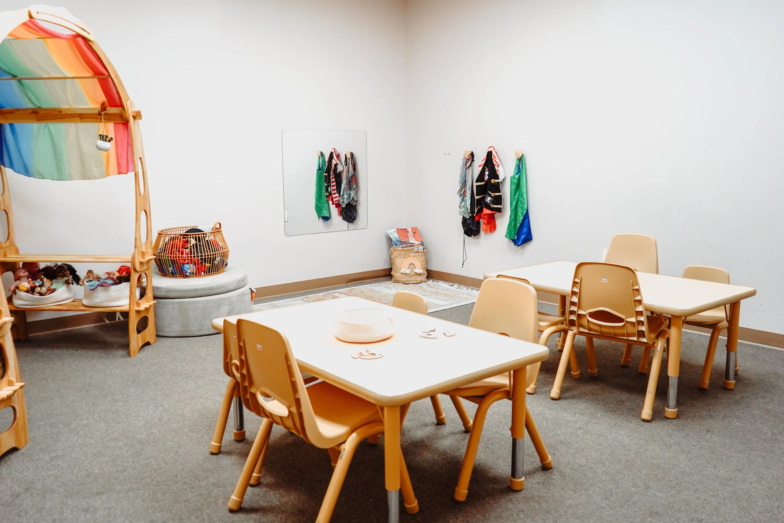 A children's playroom with beige tables and chairs, a rainbow-colored canopy, toy baskets, and costumes hanging on the wall.
