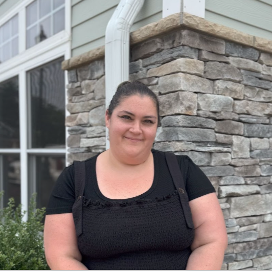 A woman with dark hair pulled back, wearing a black top, standing outside near a house with gray stone siding and large windows.