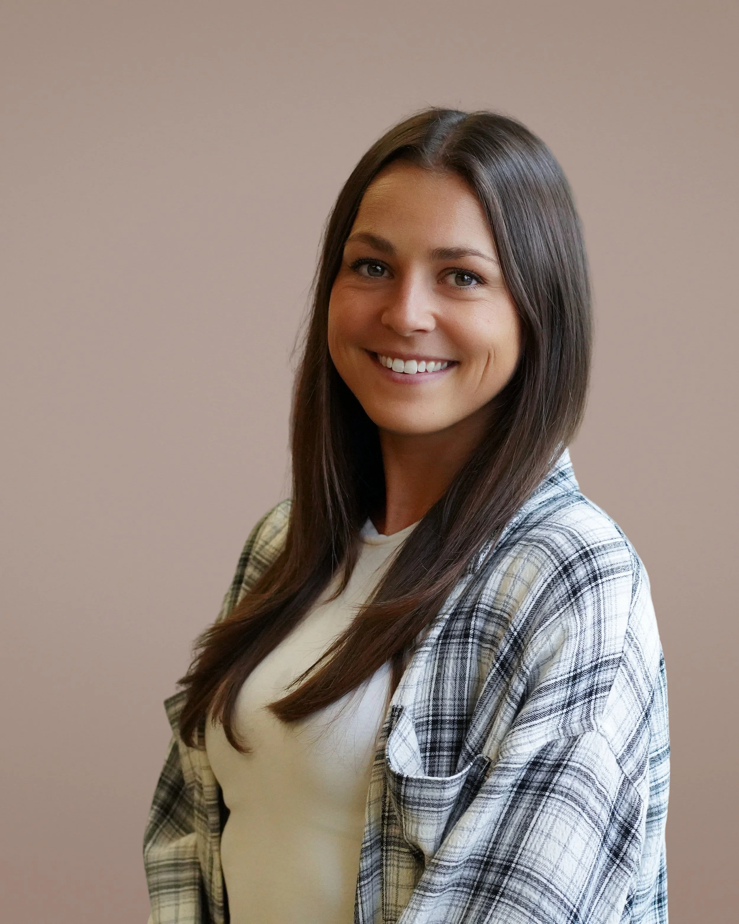 A young woman with straight brown hair, smiling, wearing a white shirt and a plaid shirt over it, standing against a plain beige wall.