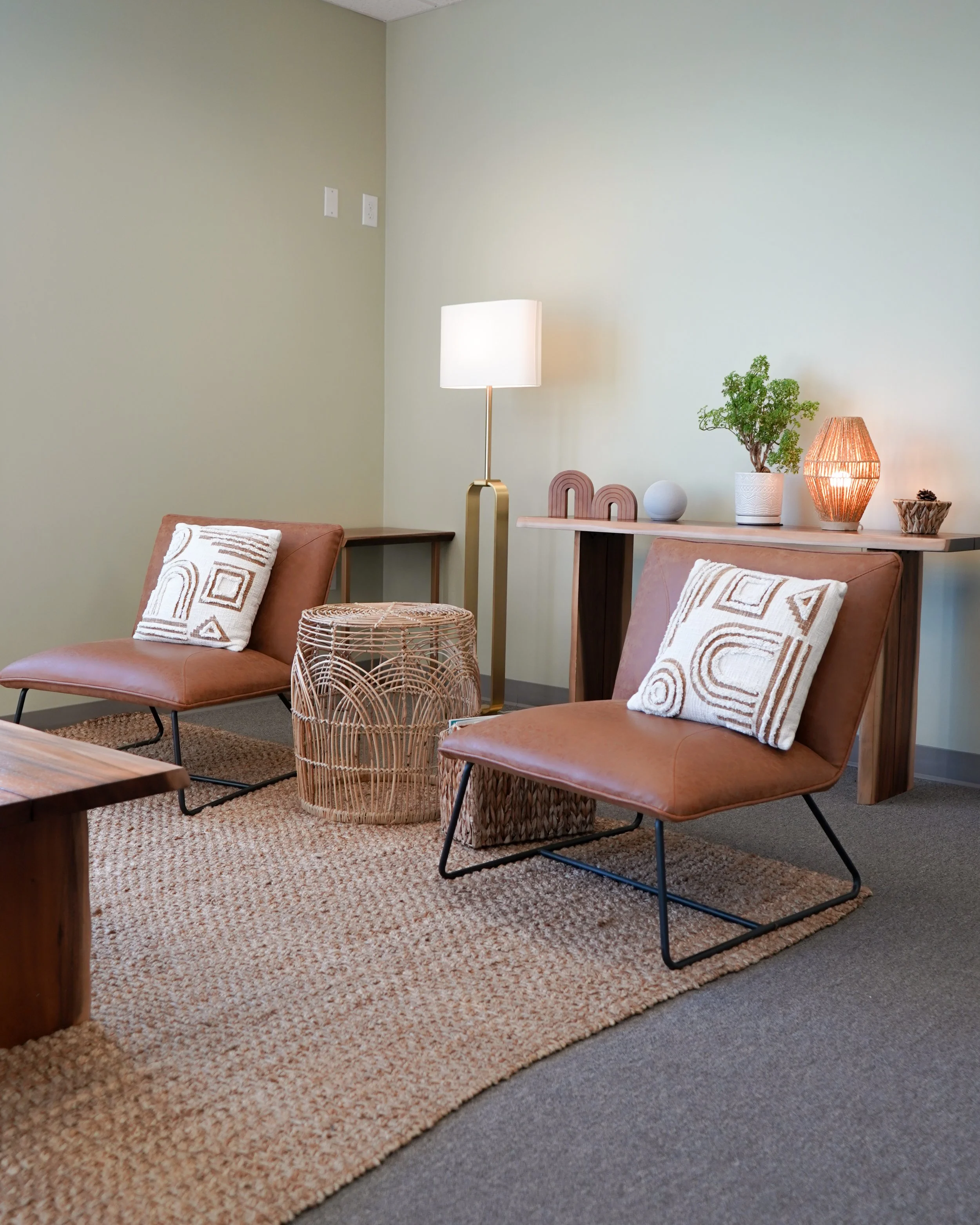 Living room with two brown leather chairs, decorative pillows, a woven side table, a wooden console table with a lamp, plant, and decorative items, on a textured rug with neutral wall color.