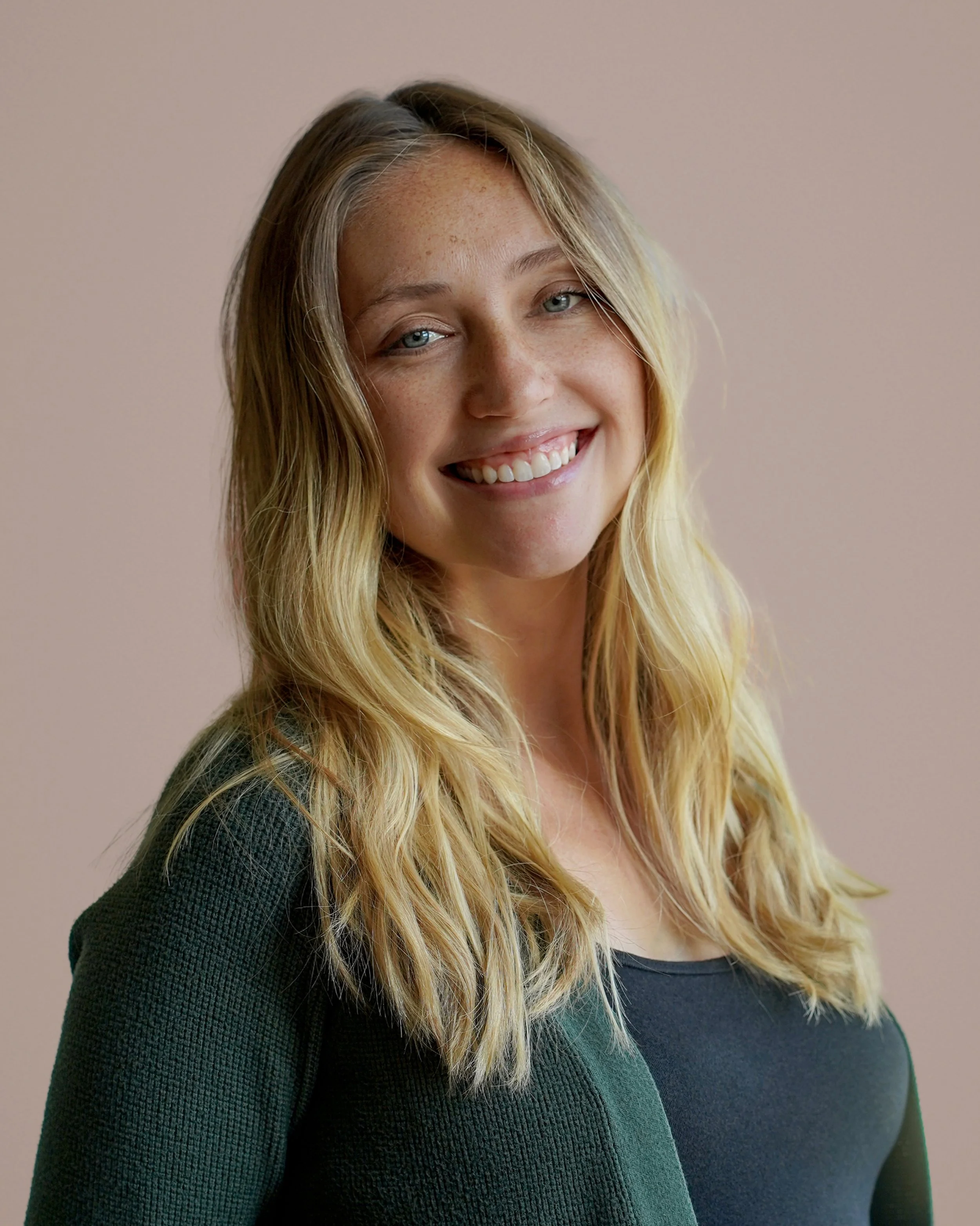 A smiling woman with long blonde hair, wearing a dark green sweater, standing against a plain pink background.