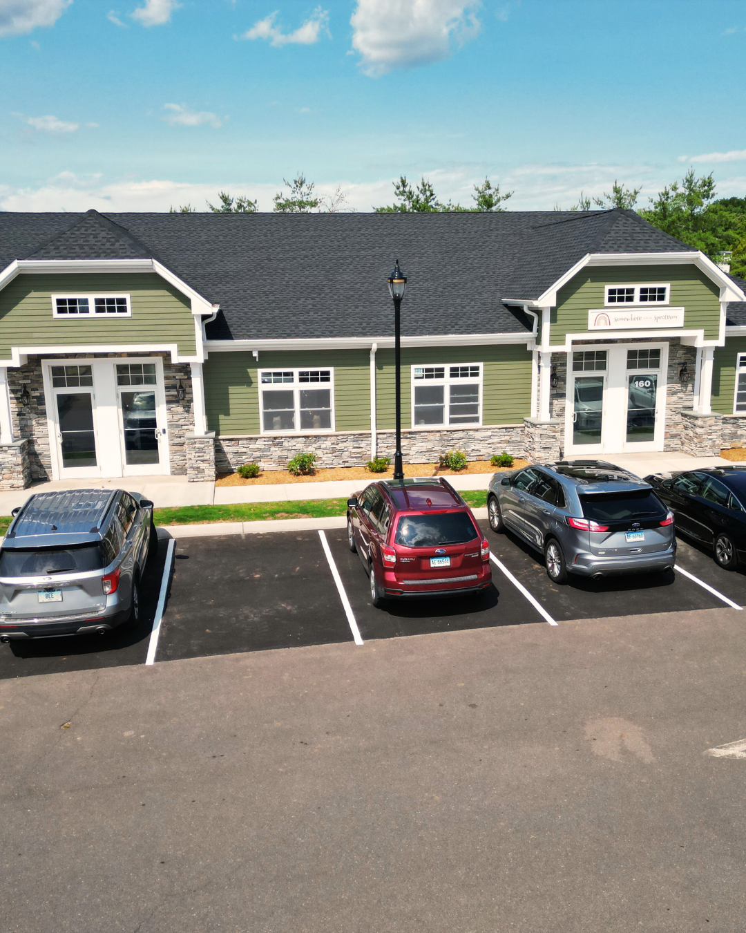 A row of parking spaces in front of a green building with stone accents, three parked cars, a black lamppost, and trees with a blue sky and clouds in the background.