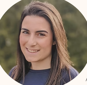 A young woman with long brown hair smiling outdoors with trees in the background.