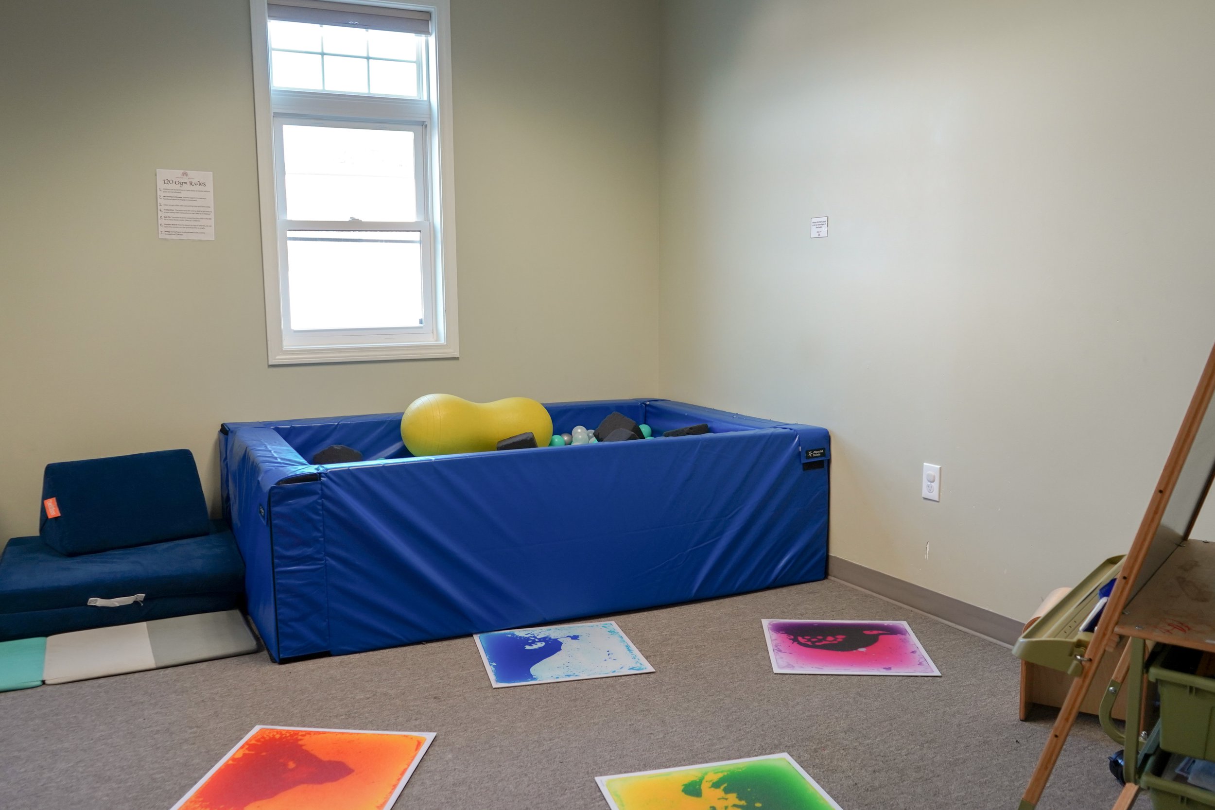 Room with a blue padded play area filled with plastic balls, a large yellow soft toy, colorful mats on the floor, a blue cushioned chair, and a window on the wall.