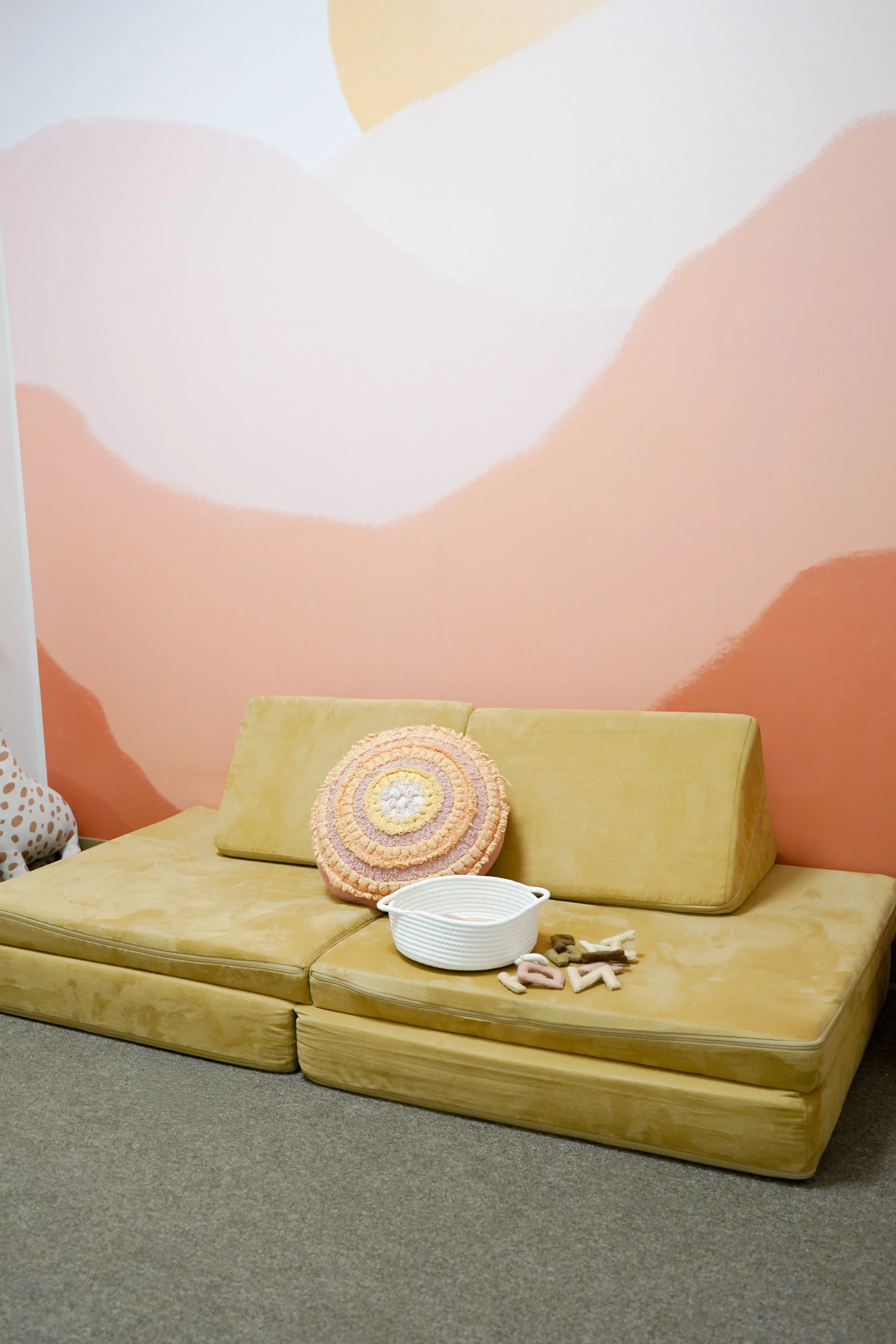 Yellow vintage sofa with a pink, yellow, and white round cushion, white basket, and felt toys on the seat, against a pastel pink and peach wall with abstract shapes.