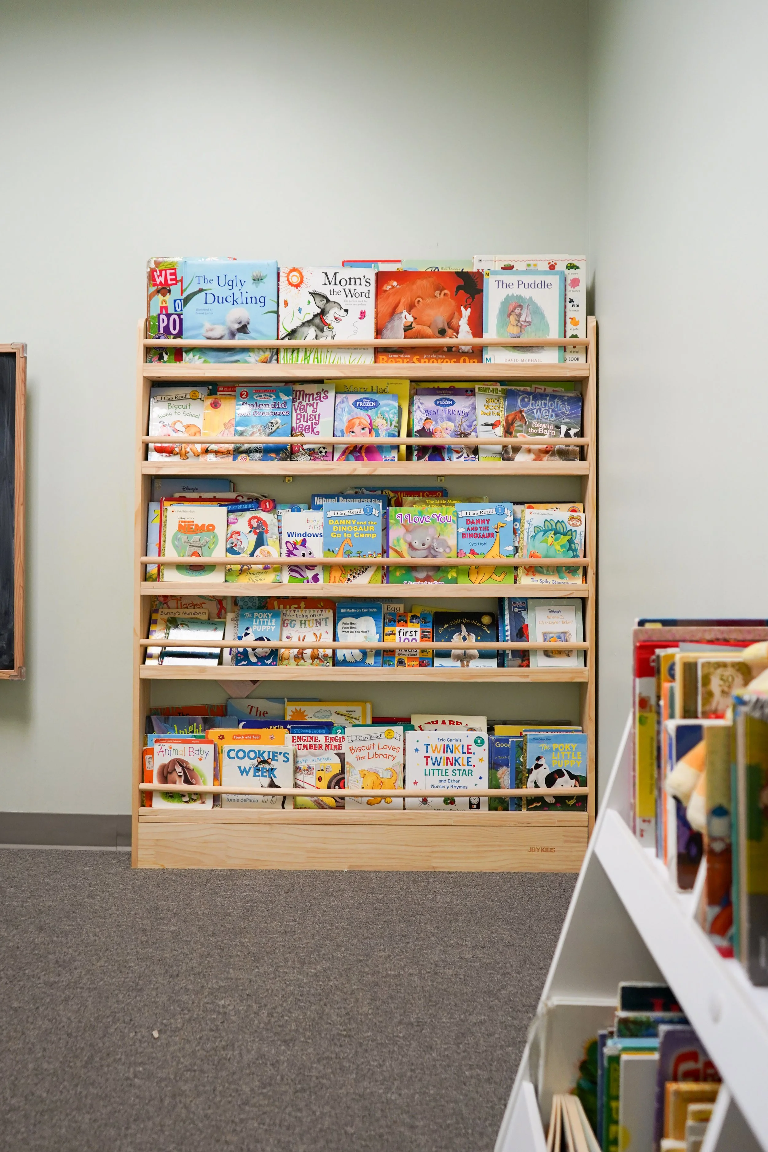 A wooden bookshelf filled with children's books in a library, with a smaller white shelf of books visible to the right.